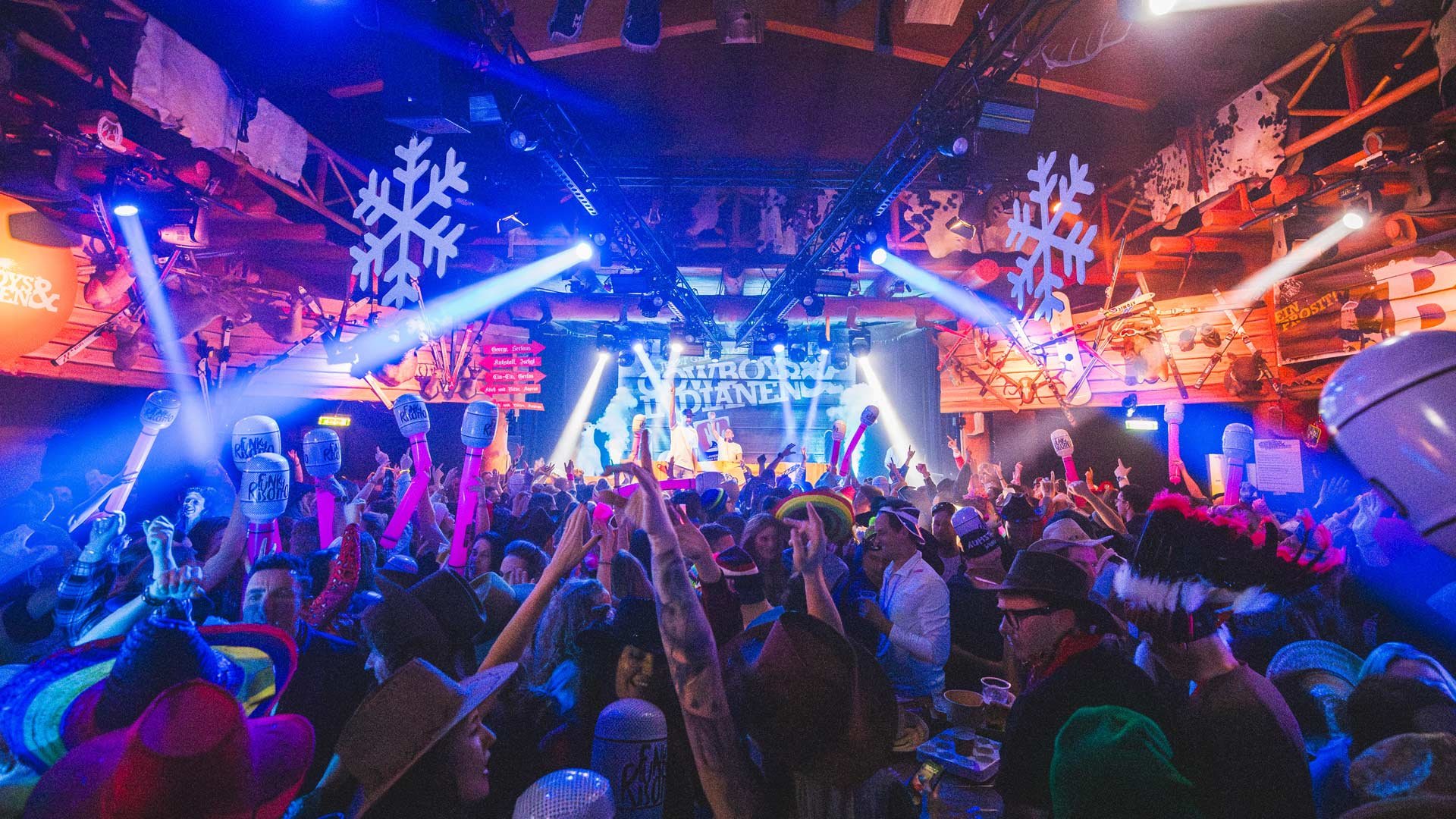 Gezellig feest met veel mensen op dansvloer, in kostuums en grappige hoeden, met feestartikelen zoals grote microfoon- en glasballonnen, op een verlichte locatie met een podium en een achtergrond die zegt "KROKUS DAGEN".