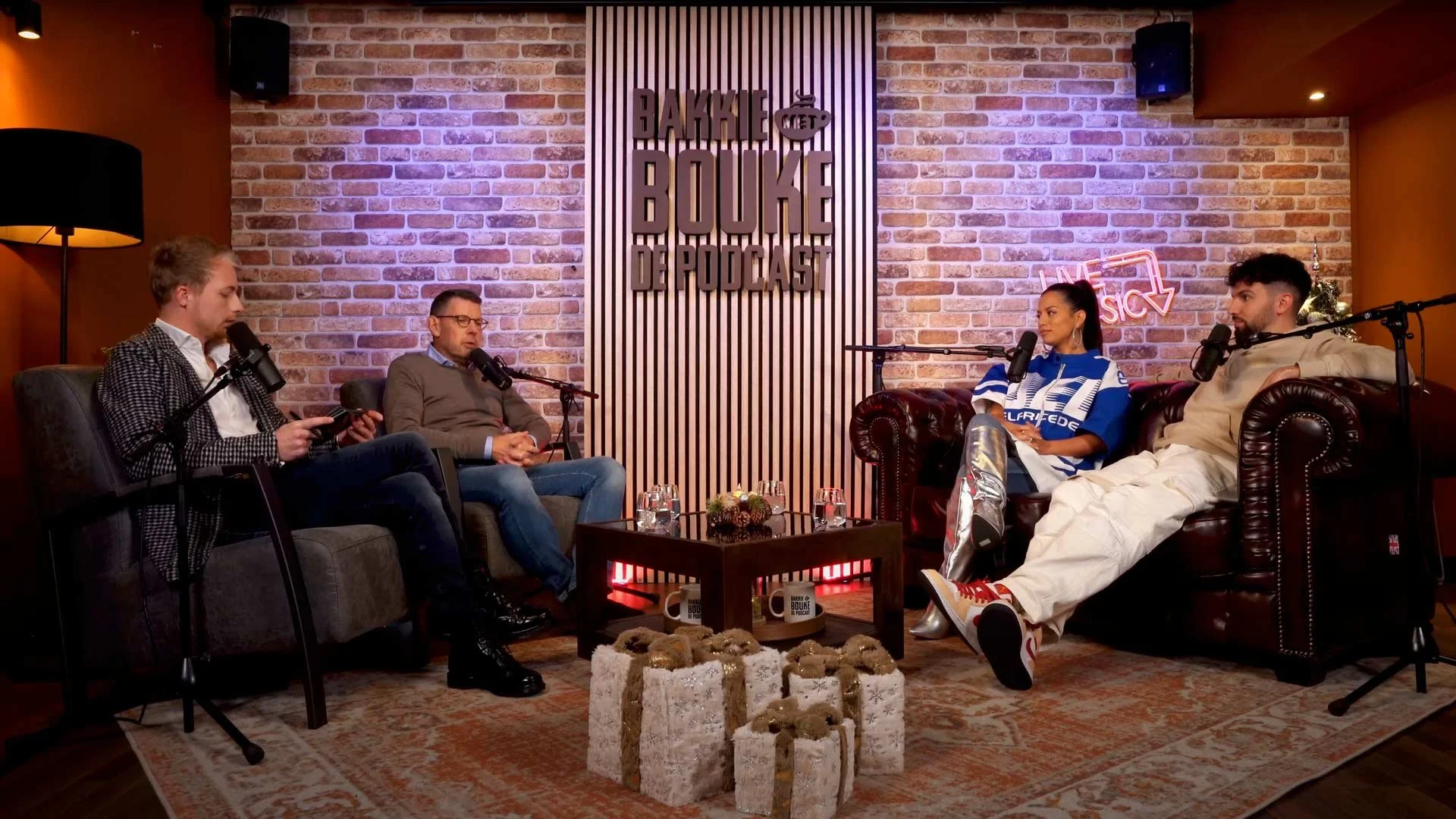 Vier mensen zitten op twee verschillende banken in een studio met een bakstenen muur, microfoons voor elk, tijdens een podcast opname.