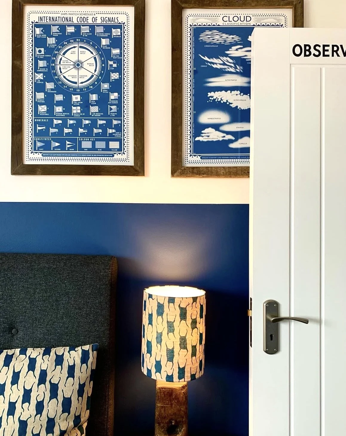 The Bluebell Inn. Wall with two framed blue posters about clouds and international signals, part of a bedside table with a patterned lampshade, a dark upholstered headboard, and a pillow with a matching pattern. 