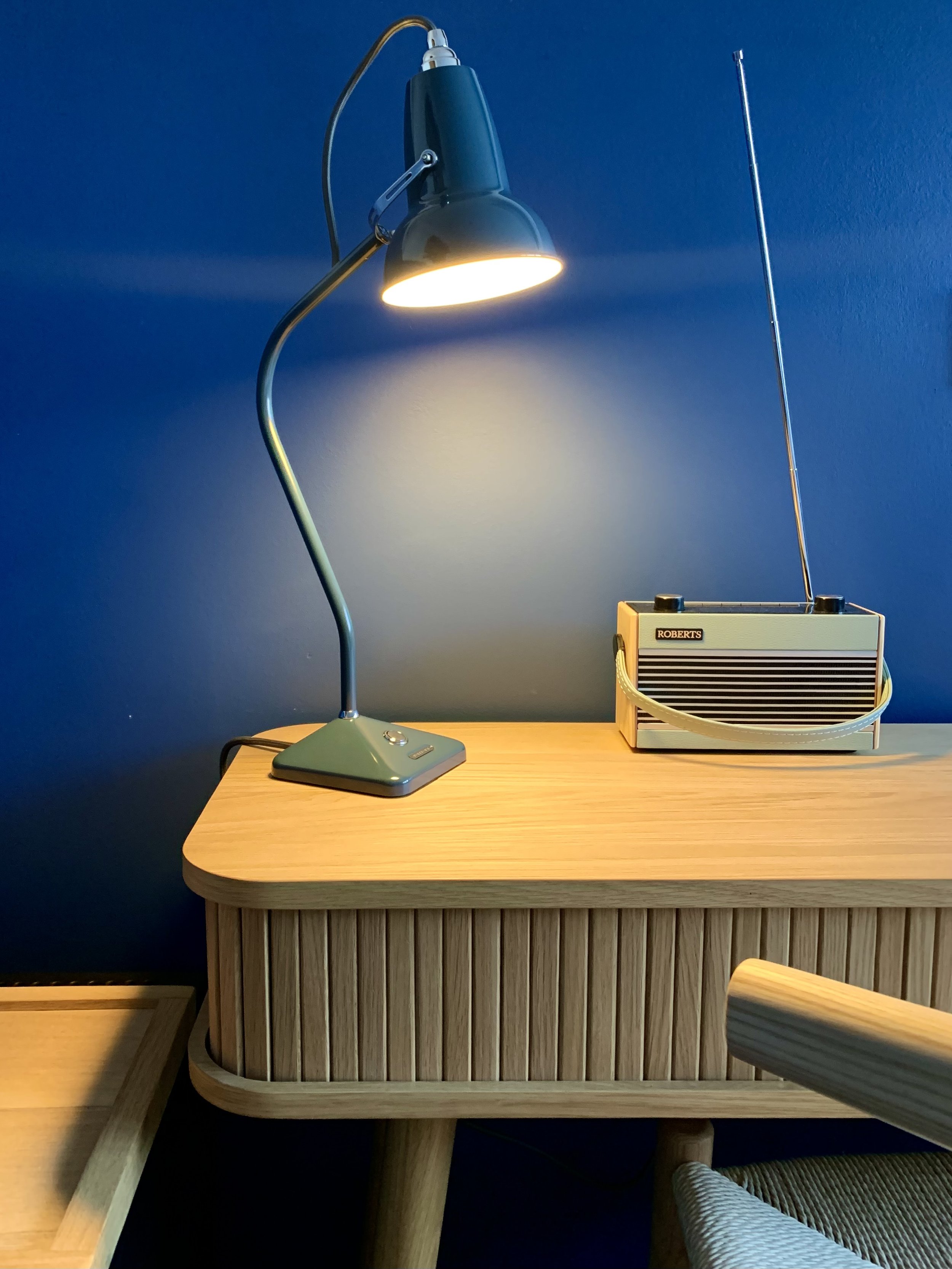 The Bluebell Inn. A blue wall with a modern table lamp turned on, positioned on a light wood table with a curved edge. To the right, there is a small vintage radio with a white handle and a silver antenna. 