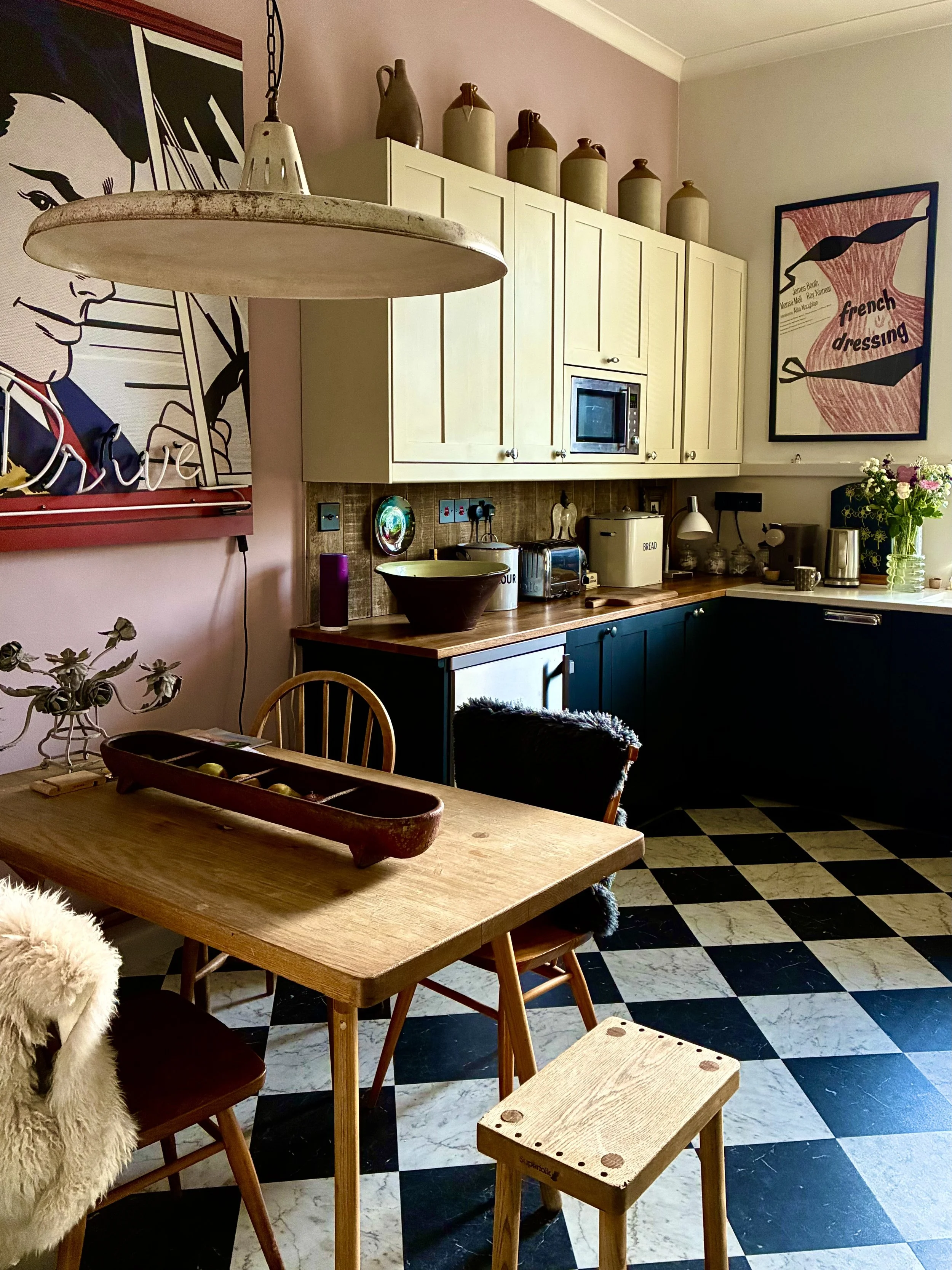 A kitchen with white cabinets, a wooden countertop, and black-and-white checkered floor. There are decorative vases on top of the cabinets, a framed artwork of a woman reading, and a poster that says 'french dressing' on the wall. The table has a lar