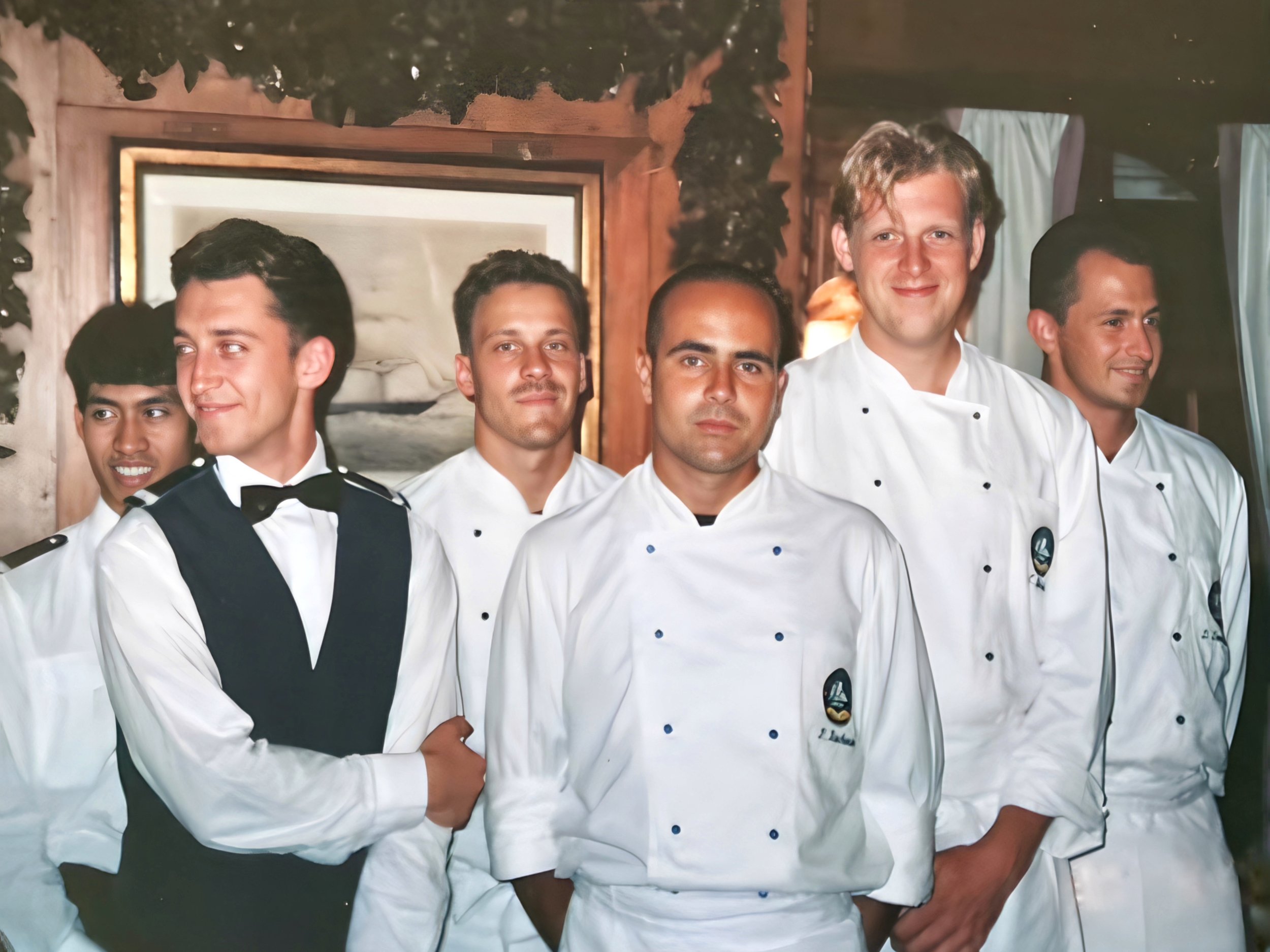Sechs Männer in Kochuniformen und ein Kellner mit Weste und Fliege stehen nebeneinander und lachen. Sie befinden sich in einem holzvertäfelten Raum.