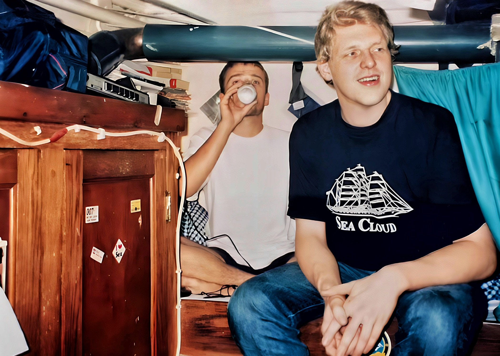 Zwei junge Männer in einer engen Kabine oder einem Wohnwagen. Einer sitzt und trinkt aus einem Becher, während der andere auf einer Treppe sitzt und ein T-Shirt mit einem Schiffsaufdruck trägt. Im Hintergrund sind Holzmöbel und persönliche Gegenständ