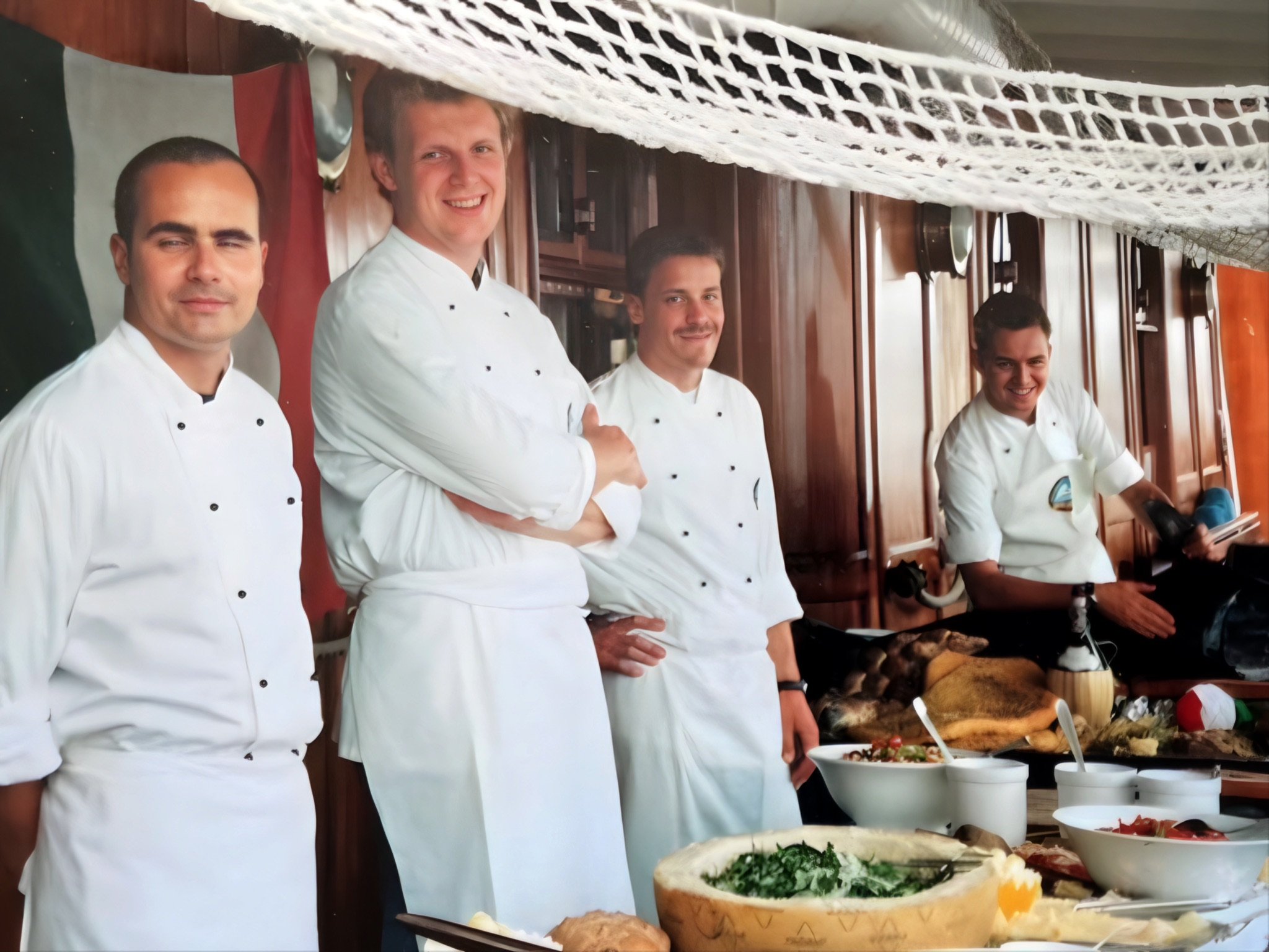 Vier Köche in weißen Kitteln lächeln in einer Restaurant- oder Küchenumgebung. Vor ihnen sind verschiedene Speisen und Zutaten auf einem Tisch ausgebreitet, darunter Käse, Salate und Brot. Im Hintergrund befindet sich eine italienische Flagge.