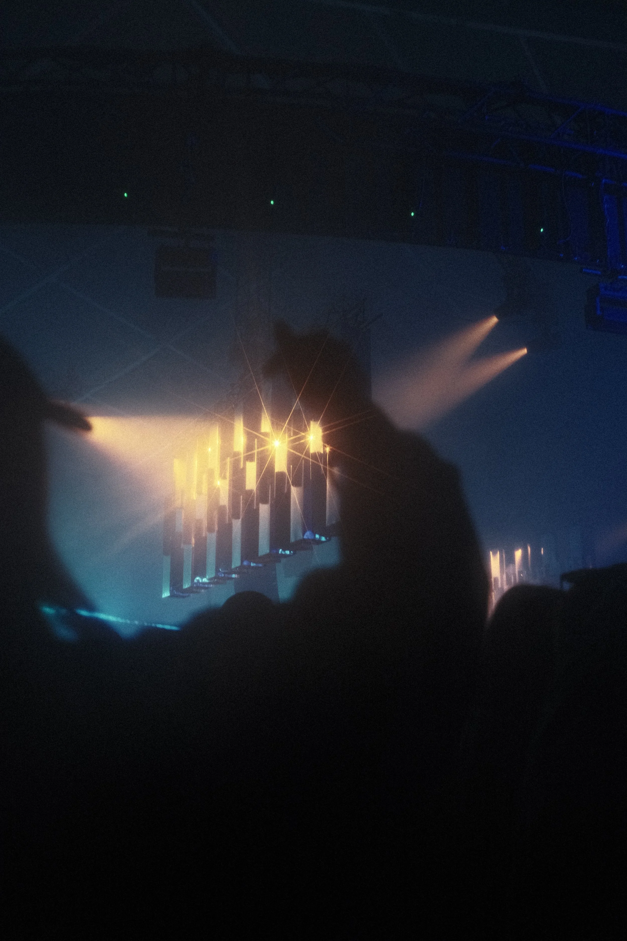 Concert or nightclub scene with stage lighting, projection on the wall, and silhouettes of concert attendees in the foreground. Reflecting Bodies VI Voodoo Village Belgium Scenography art installation light installation Simone Smelt Lumus Instruments