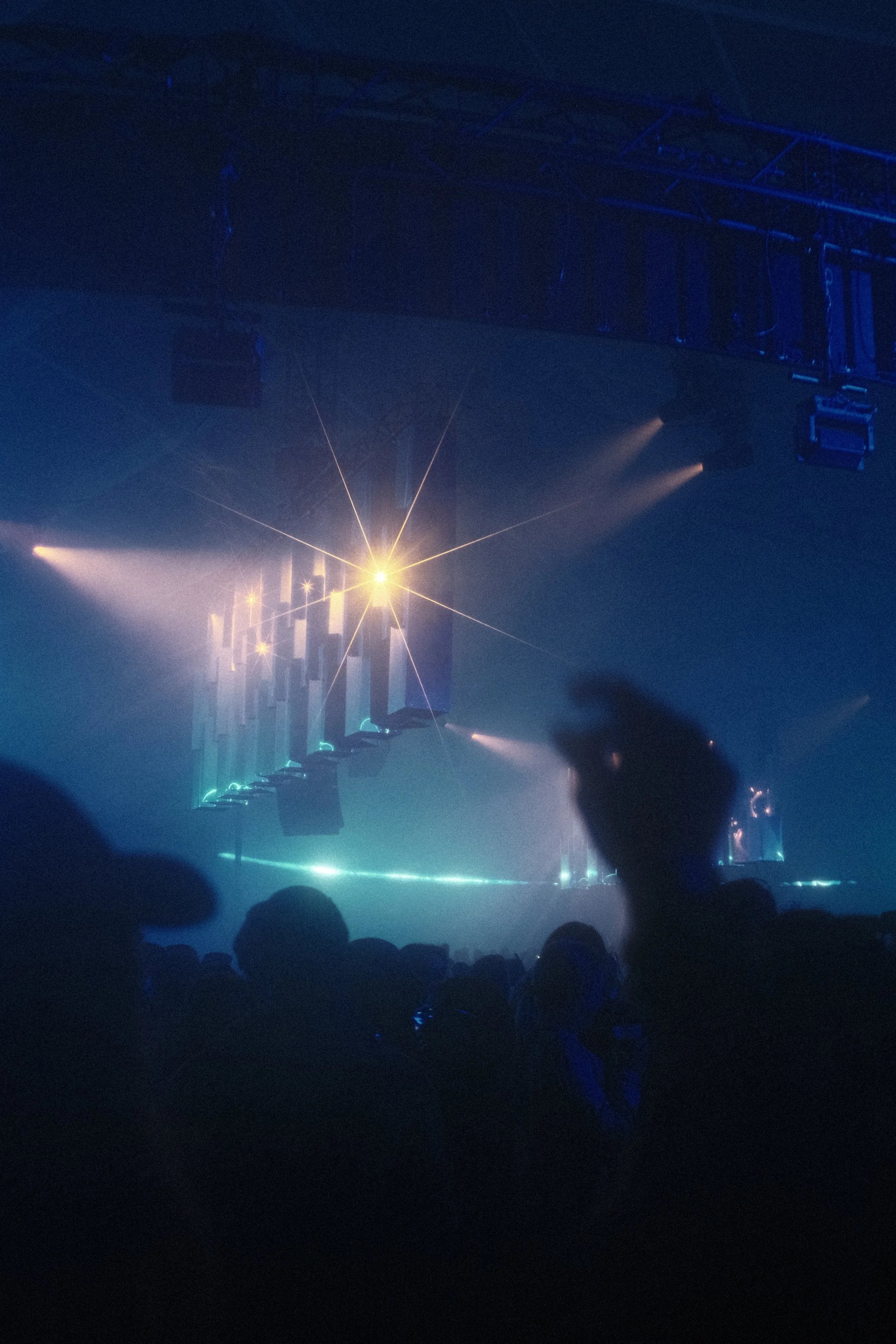 A concert scene with stage lights and spotlights illuminating a dark venue, with silhouettes of audience members in the foreground. Reflecting Bodies VI Voodoo Village Belgium Scenography art installation light installation Simone Smelt Lumus Instrum