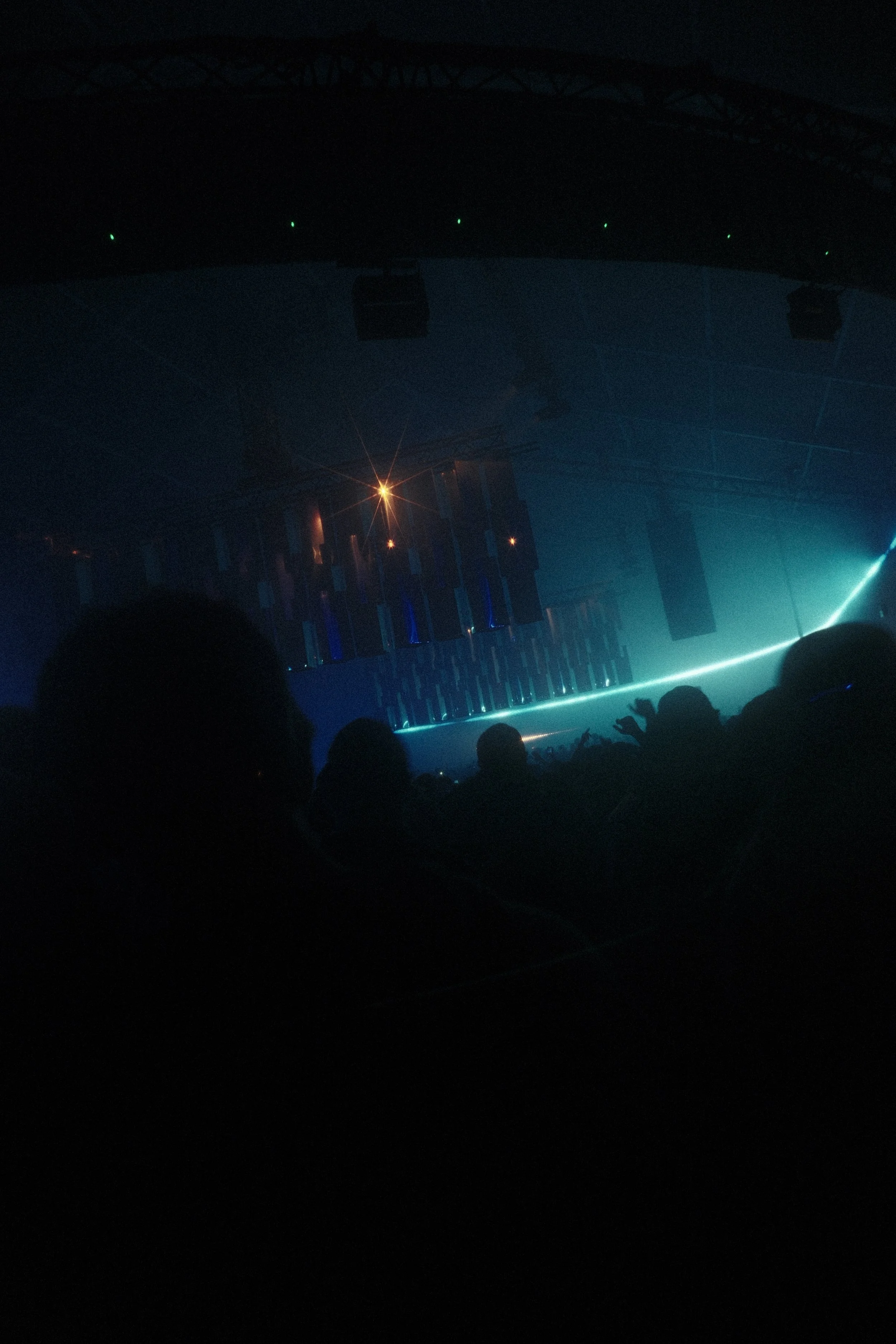 Dark concert venue with dim lighting, audience silhouette, stage with blue lighting, and hanging speakers. Reflecting Bodies VI Voodoo Village Belgium Scenography art installation light installation Simone Smelt Lumus Instruments Festival main stage