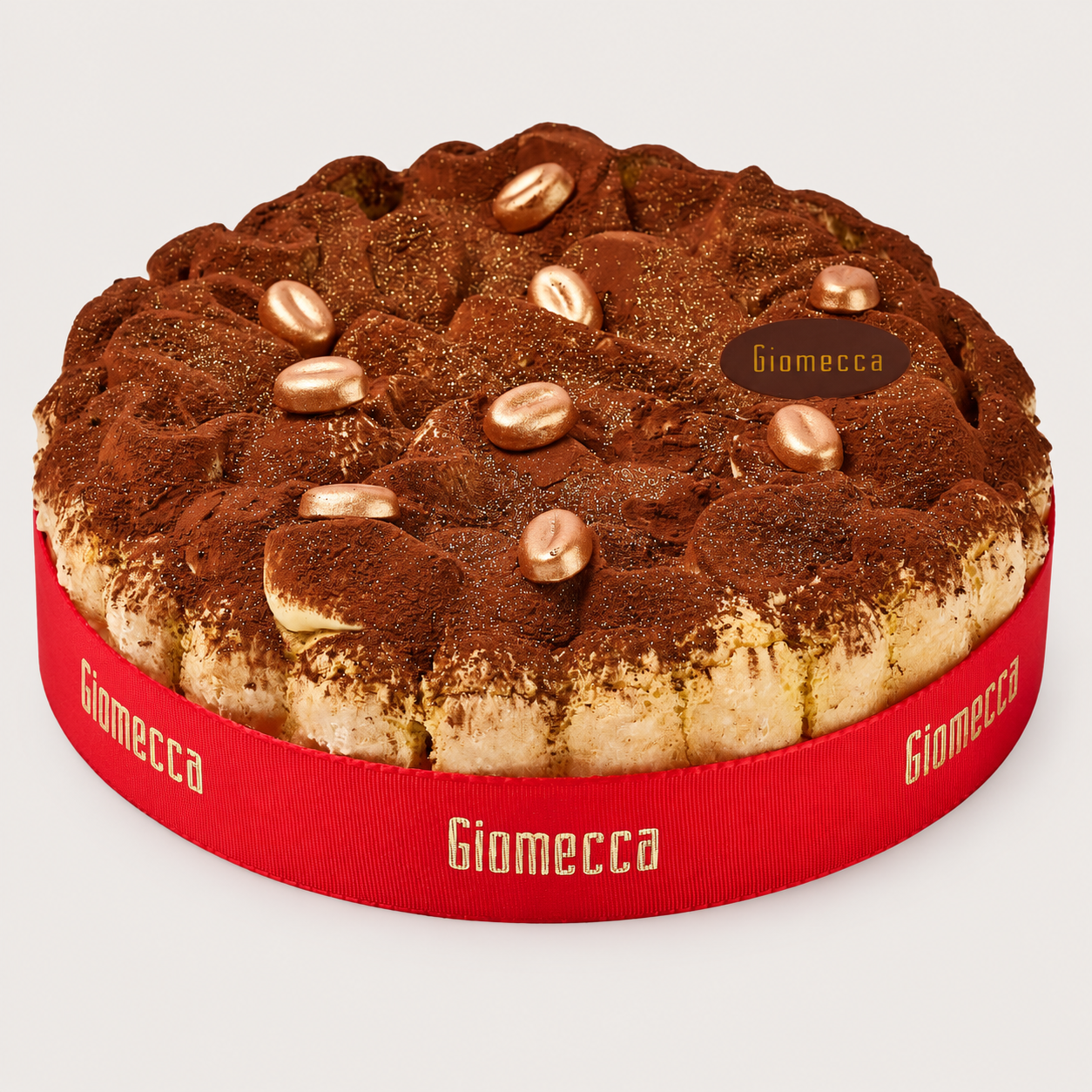 Round vanilla cake topped with chocolate and gold candies, wrapped with a red ribbon that says 'Giomecca.'
