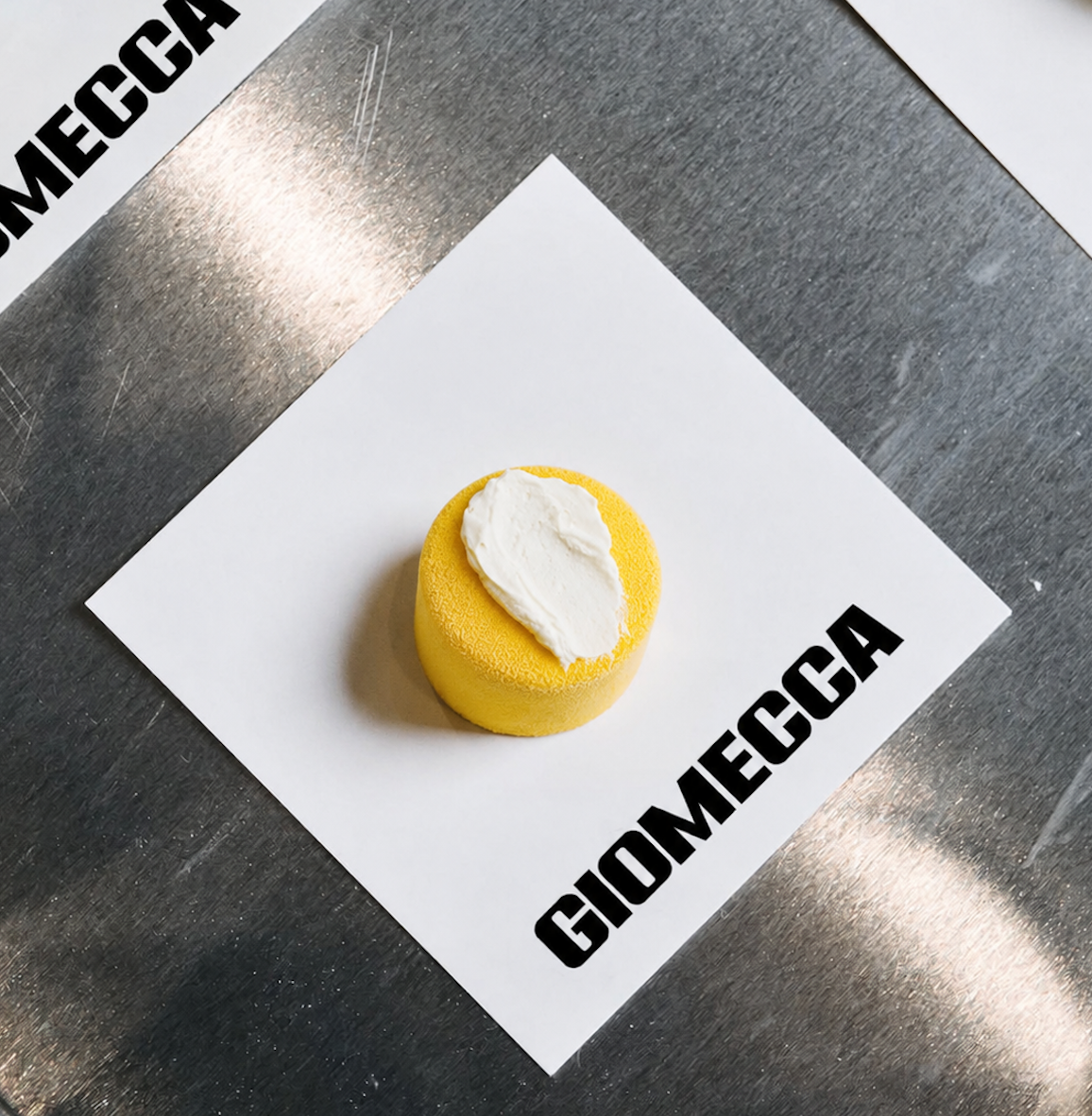 A yellow, round cake with a white cream topping placed on a white sheet of paper with black text, on a metallic surface.