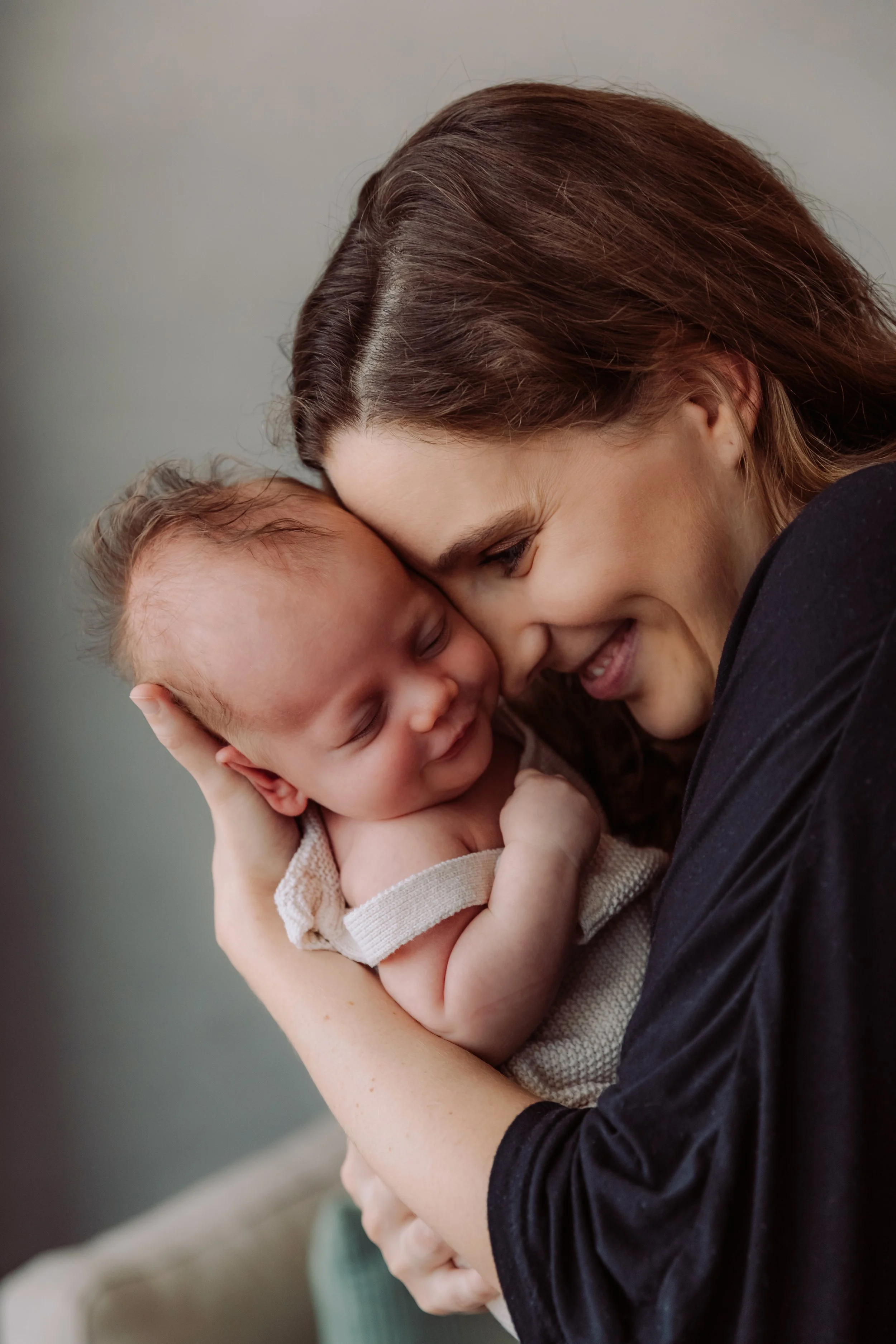 A woman is holding a sleeping baby close, touching foreheads, both smiling, in a tender moment.