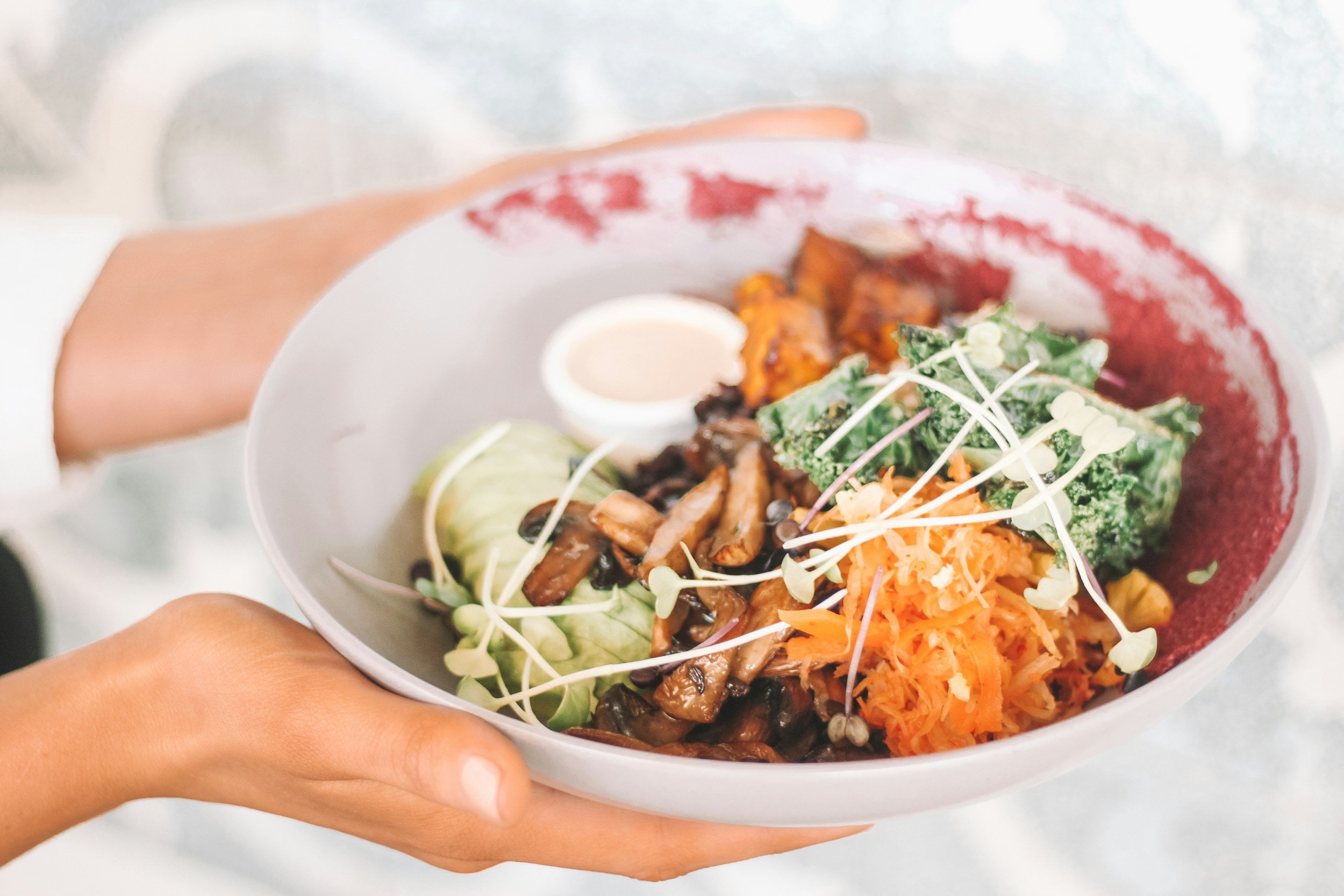 A person holding a white bowl of mixed salad with shredded carrots, kale, microgreens, sliced cucumber, grilled mushrooms, and a small cup of dressing.