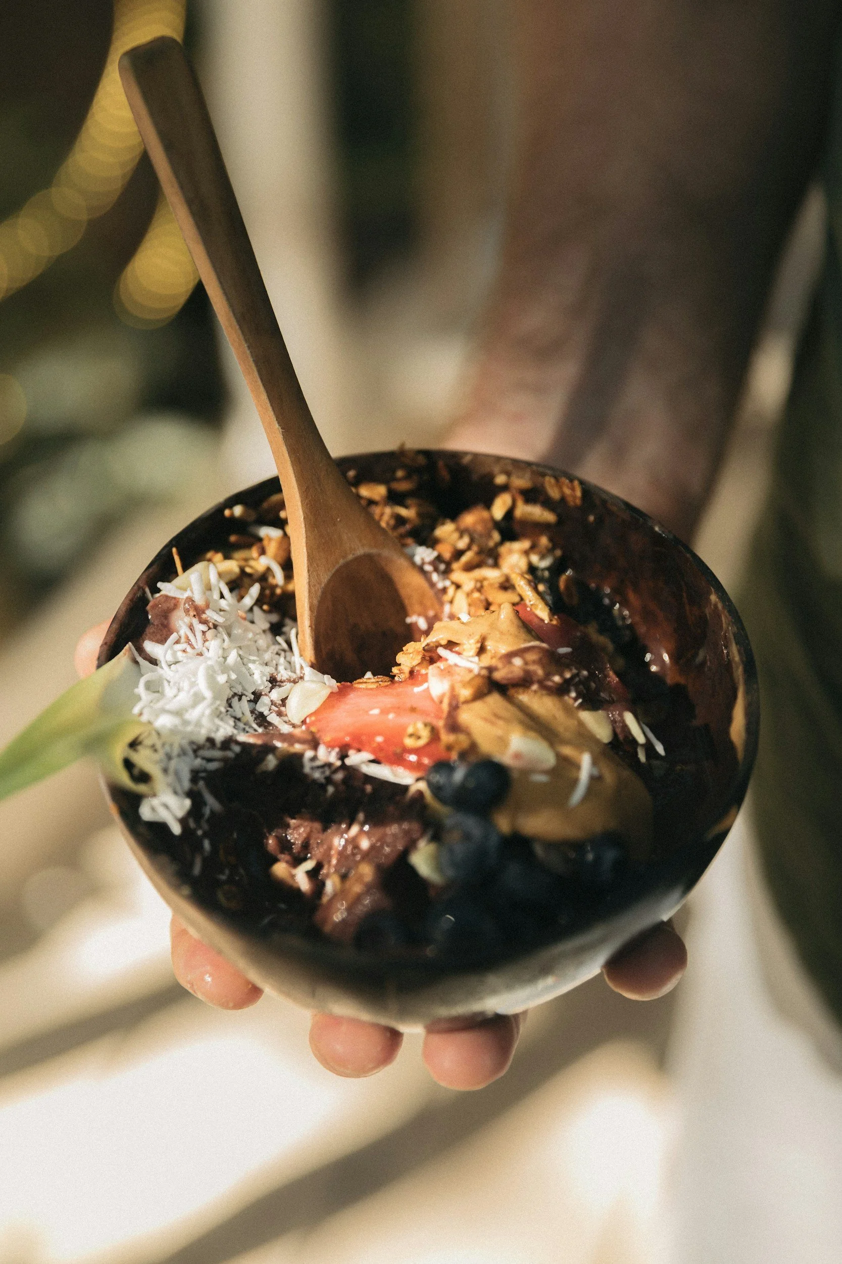 A hand holding a bowl of ice cream topped with chocolate, berries, shredded coconut, nuts, and caramel, with a wooden spoon inside.