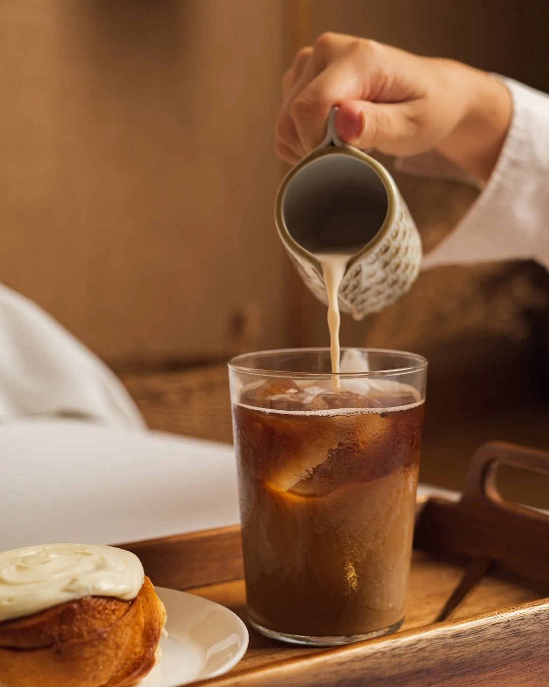 A hand pouring cream into a glass of iced coffee on a wooden tray, with a plate of cinnamon roll with white icing nearby.