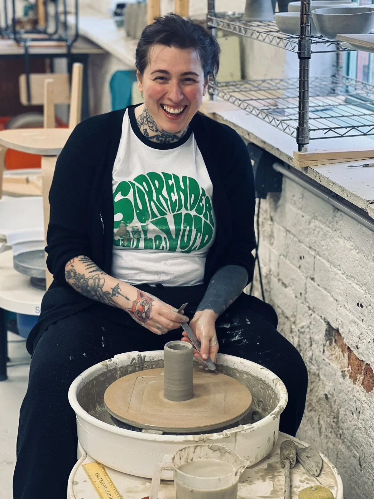 A woman with tattoos on her arms and neck is smiling while working on a pottery wheel, shaping a gray clay vessel in a ceramic studio.