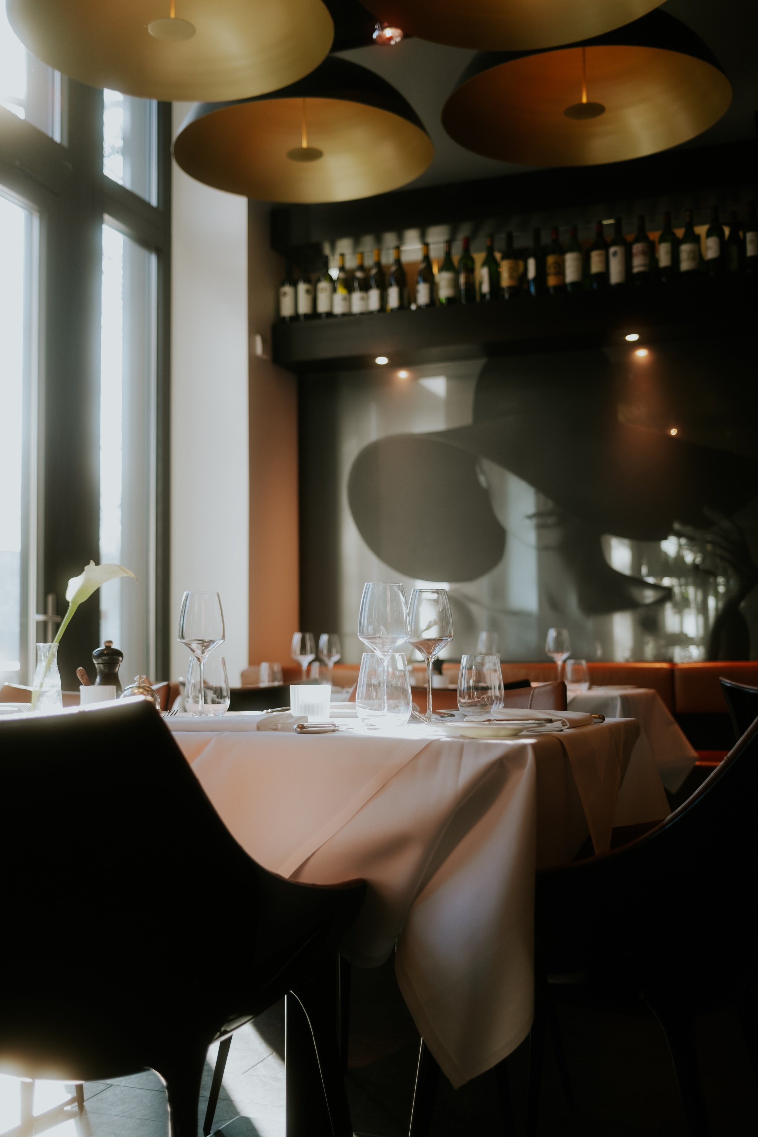 Elegantes Restaurant mit gedecktem Tisch, Weingläsern und Dekorflasche, im Hintergrund Weinflaschen und stilvolles Porträt einer Person mit großem Hut.
