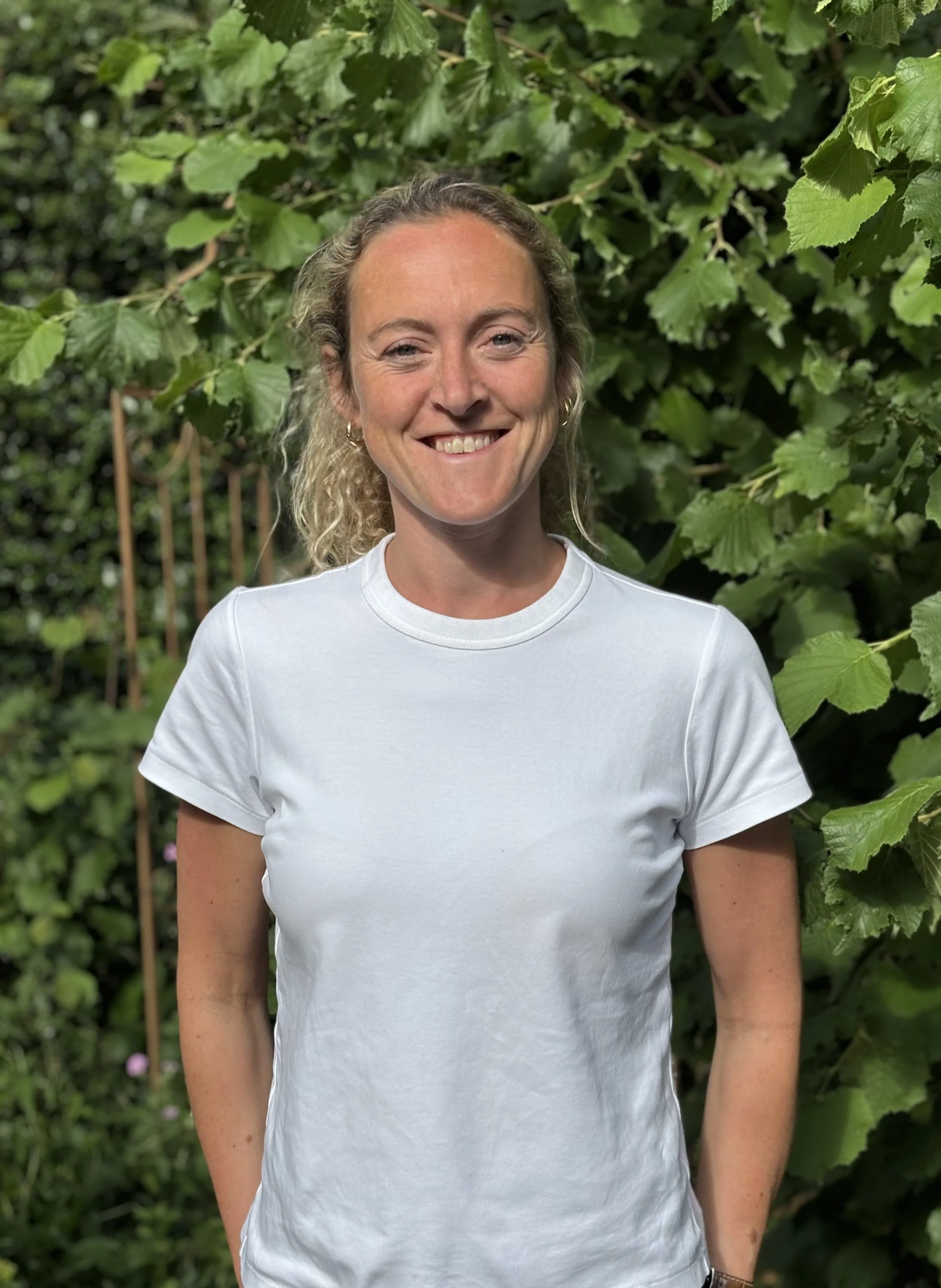 A woman with curly blonde hair, wearing a white t-shirt, smiling and standing outdoors in front of green leafy plants.