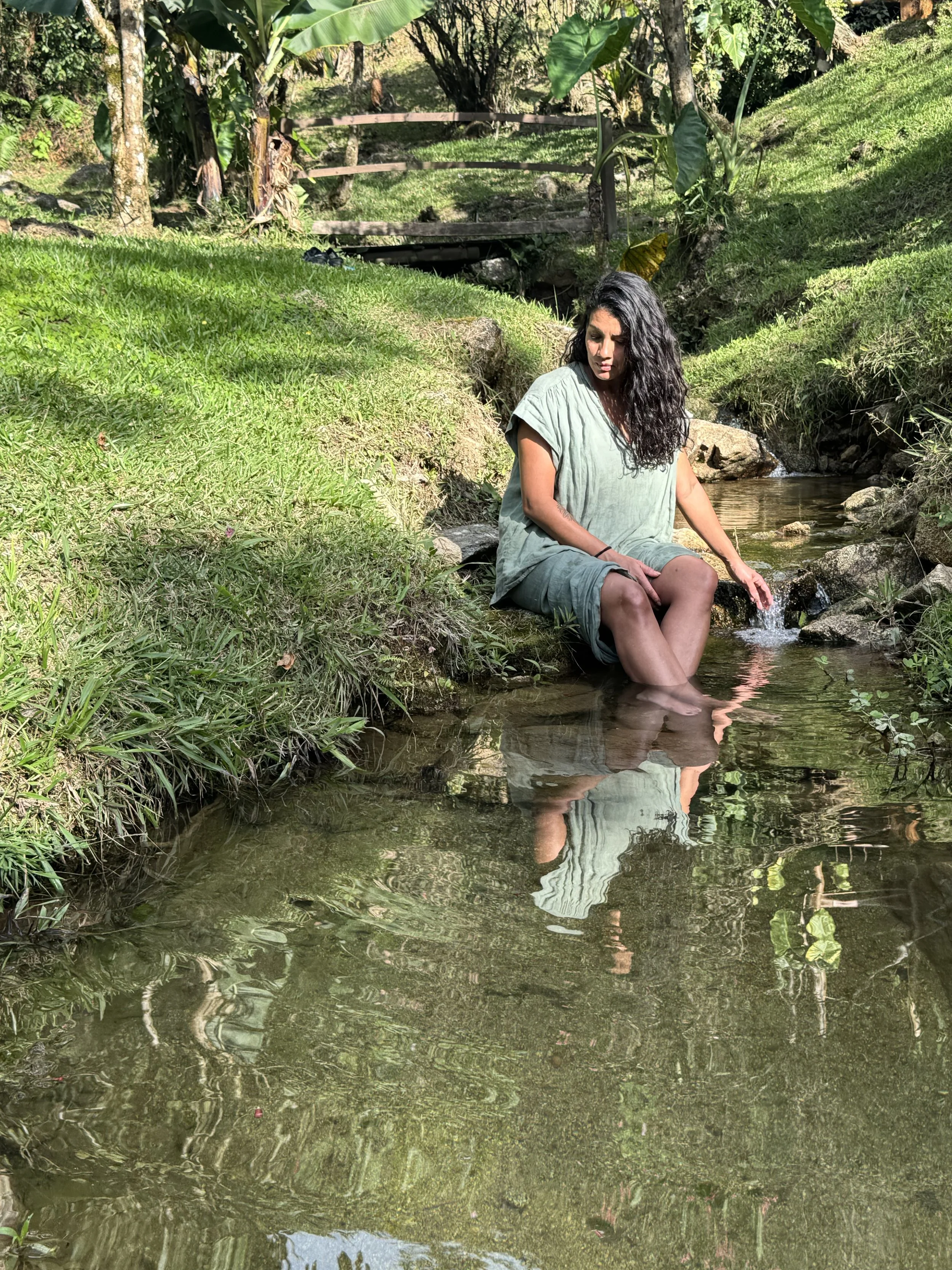 En kvinne som sitter på kanten av en liten bekk i en grønn hage med store blader og trær, og reflekteres i vannet.