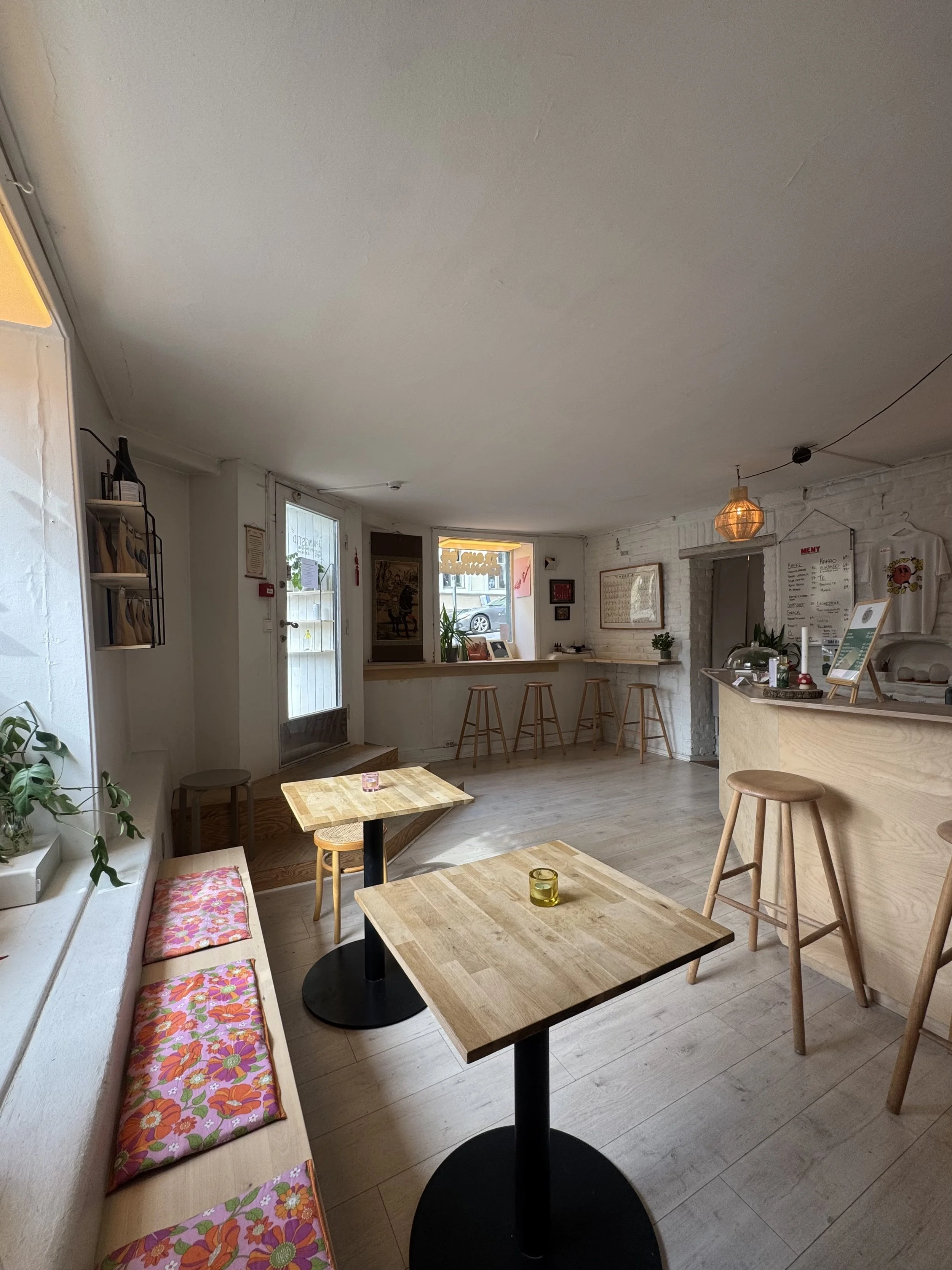 Interiørbilde av en koselig café med trebord, barstoler, blomster, og en liten bar i bakgrunnen med en meny. Naturlig lys fra vinduer, lys treverk og rolig atmosfære.