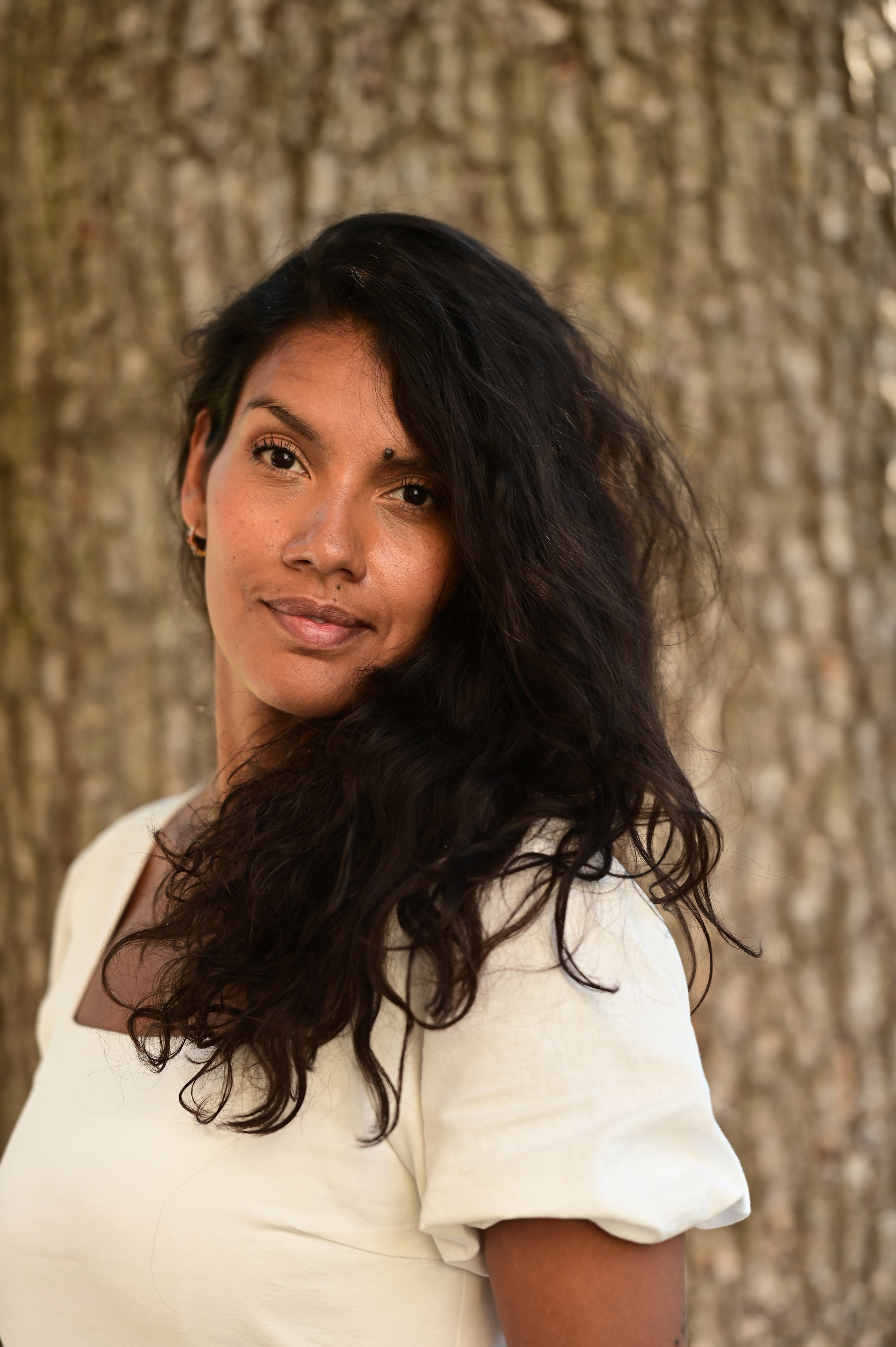 Et portrett av en kvinne med mørkt, krøllete hår og perleører, stående foran et tre med bark. Angela Johansson