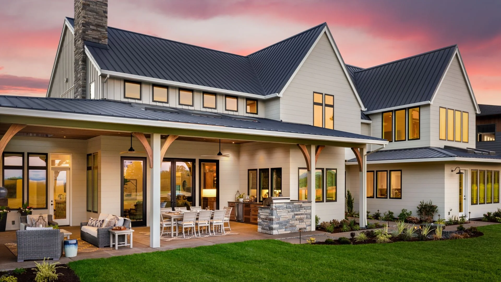 A modern two-story house featuring a covered patio with outdoor seating, a stone fireplace, and lush landscaped yard at sunset.