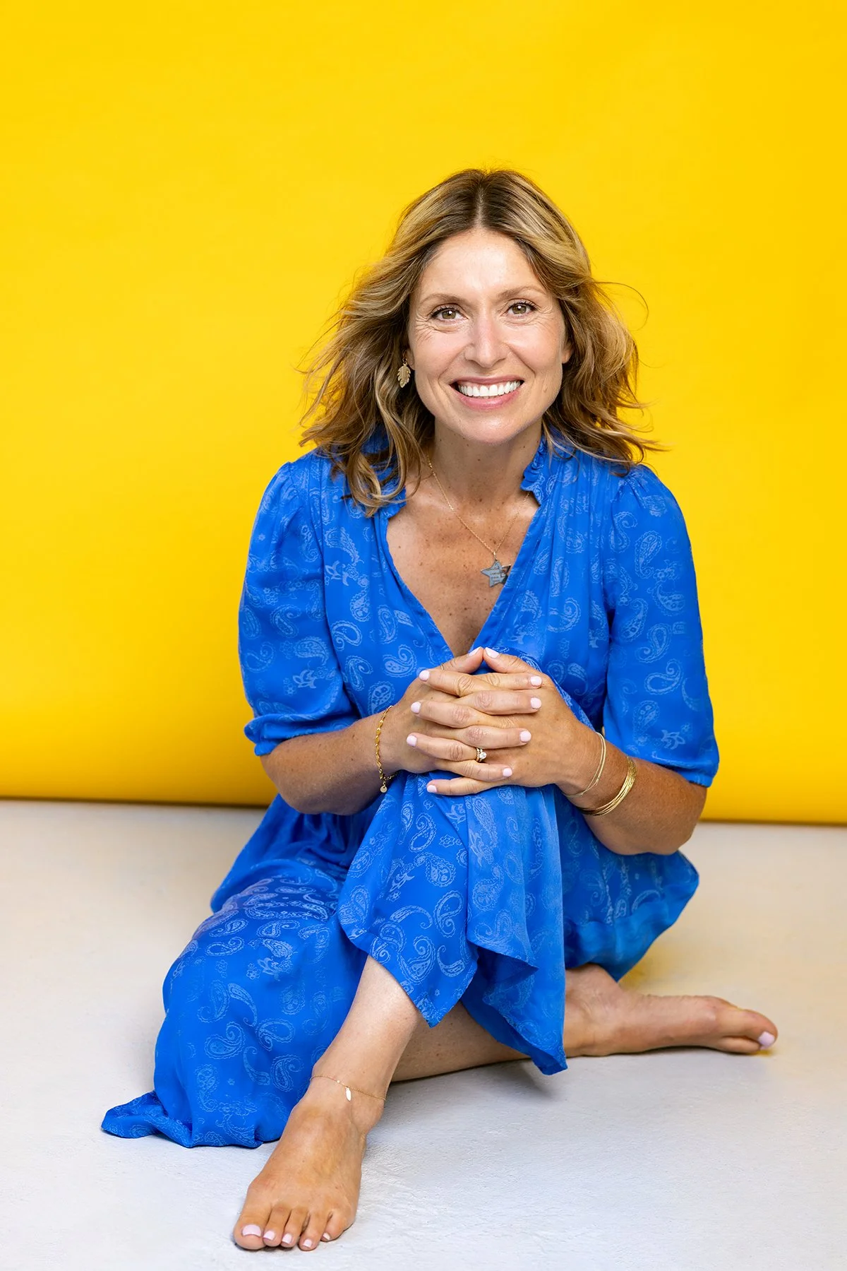 A smiling woman with shoulder-length wavy hair sitting on the floor with a yellow background behind her, wearing a blue dress and gold jewelry.