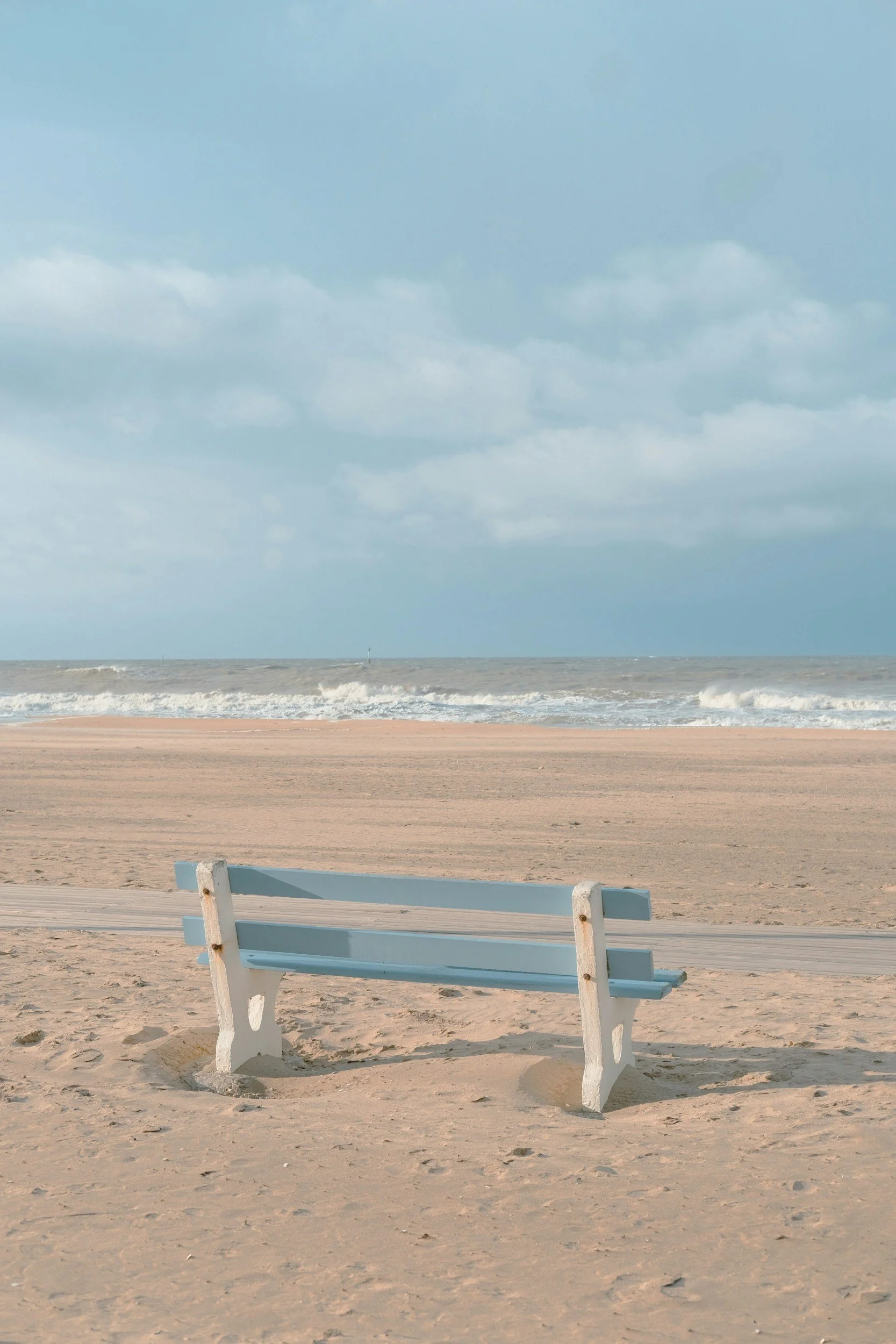 Empty light blue bench on sandy beach with ocean waves in the background and partly cloudy sky.