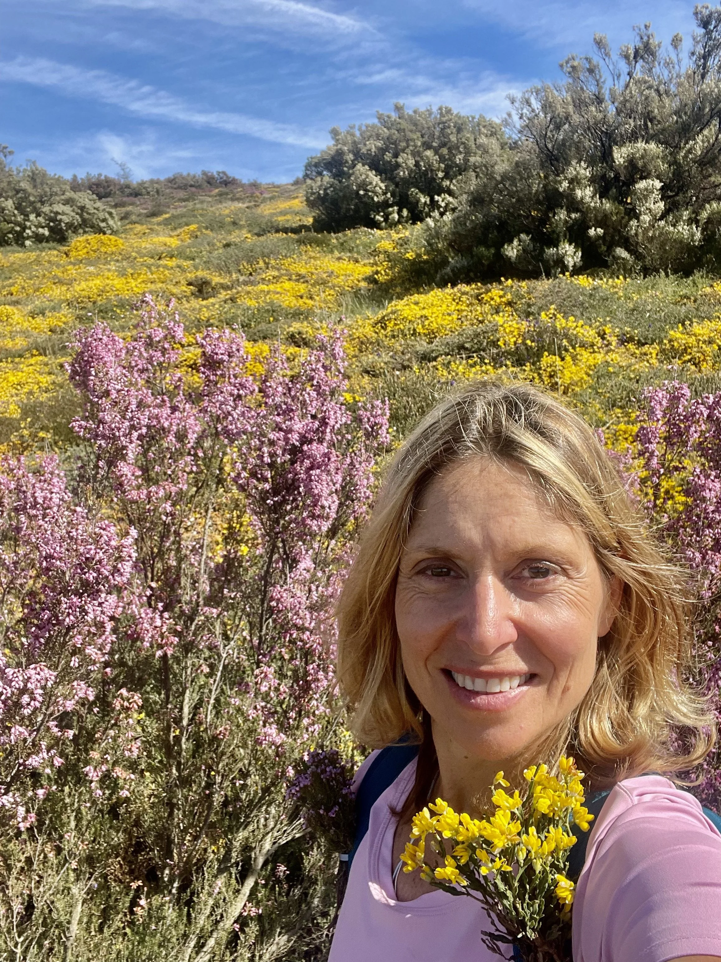 Une femme paisible et souriante aux cheveux blonds, portant une chemise rose pâle et un sac à dos, prenant un selfie dans un champ de fleurs sauvages roses et jaunes par une journée ensoleillée, avec un ciel bleu et des collines en arrière-plan.