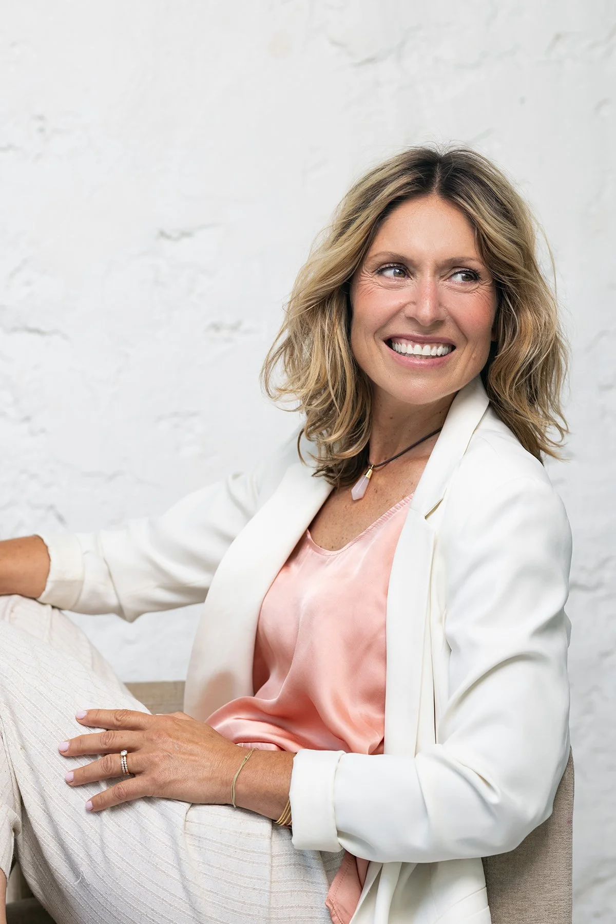 A woman with shoulder-length blonde hair, smiling, wearing a light pink satin top, white blazer, and beige pants, sitting against a white textured wall.