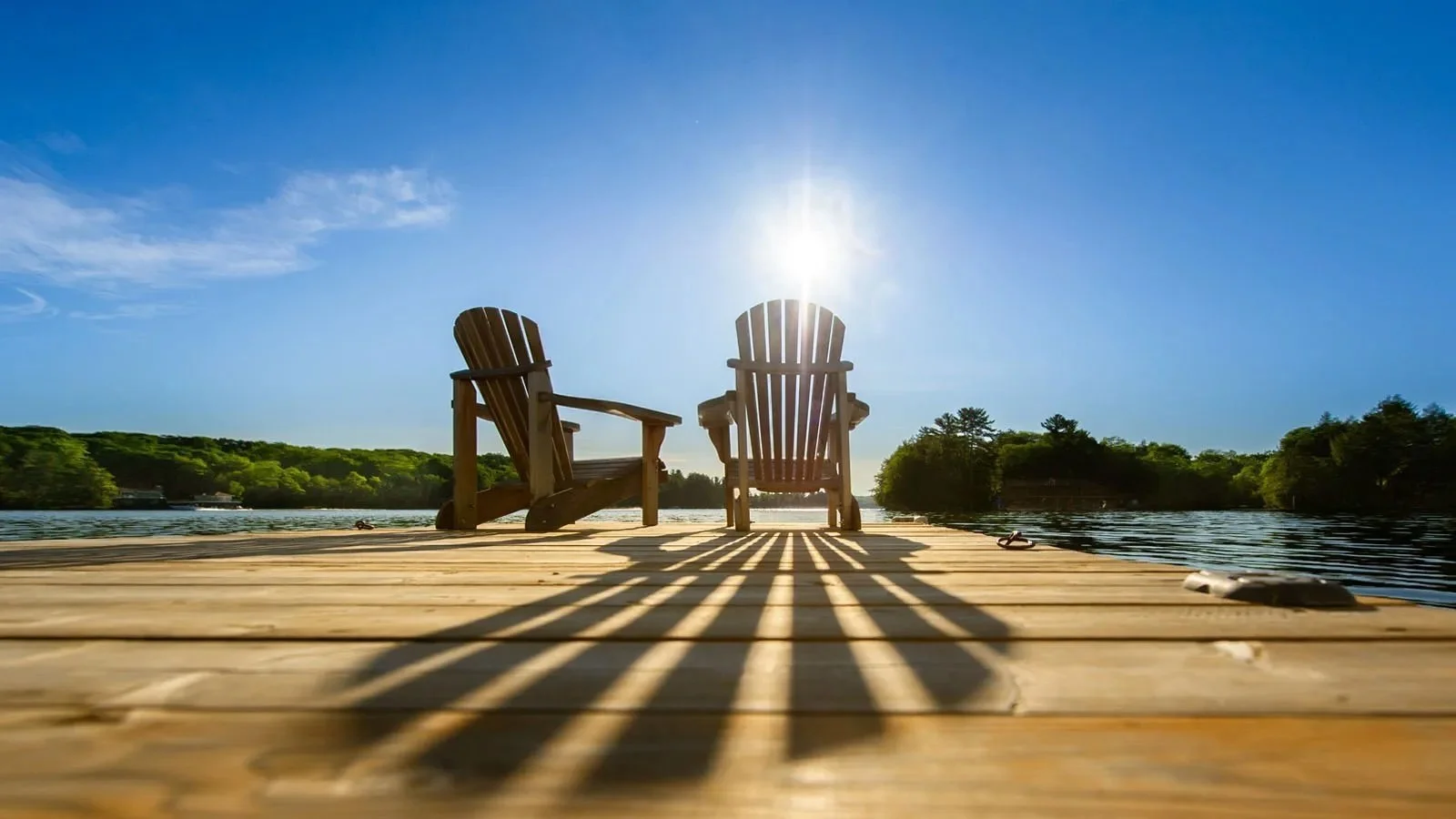 Two wooden Adirondack chairs on a dock overlooking a lake with trees in the background under a bright, clear sky.