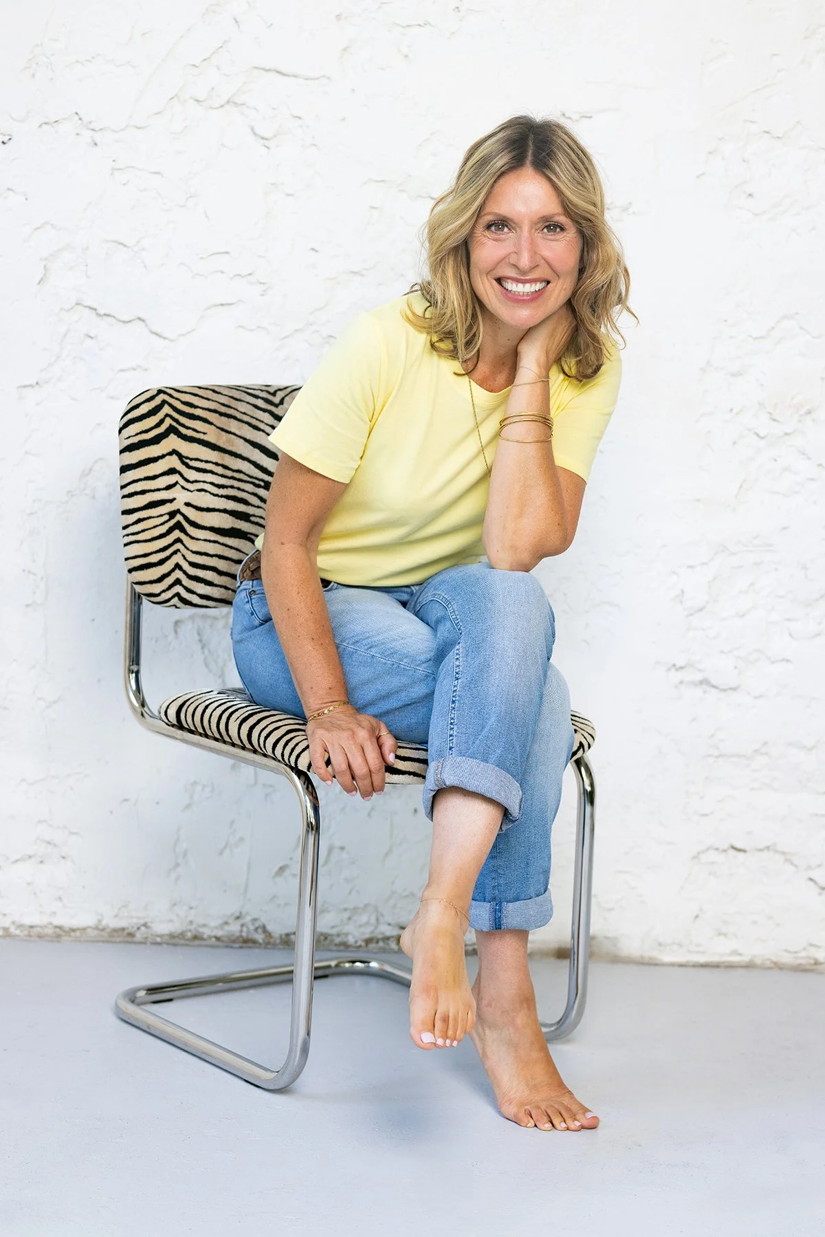 A woman with blonde, curly hair, smiling, sitting on a chair with zebra print upholstery, wearing a yellow t-shirt, cuffed blue jeans, and barefoot, against a white textured wall.