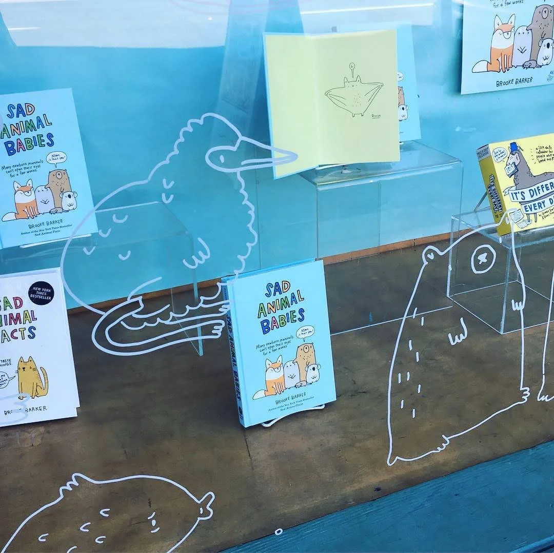 Bookstore window display featuring books titled 'Sad Animal Babies' with cartoon animal illustrations, including a bird, a frog, and a bear, and a yellow book with similar artwork. Decorative animal outlines and a blue background enhance the display.