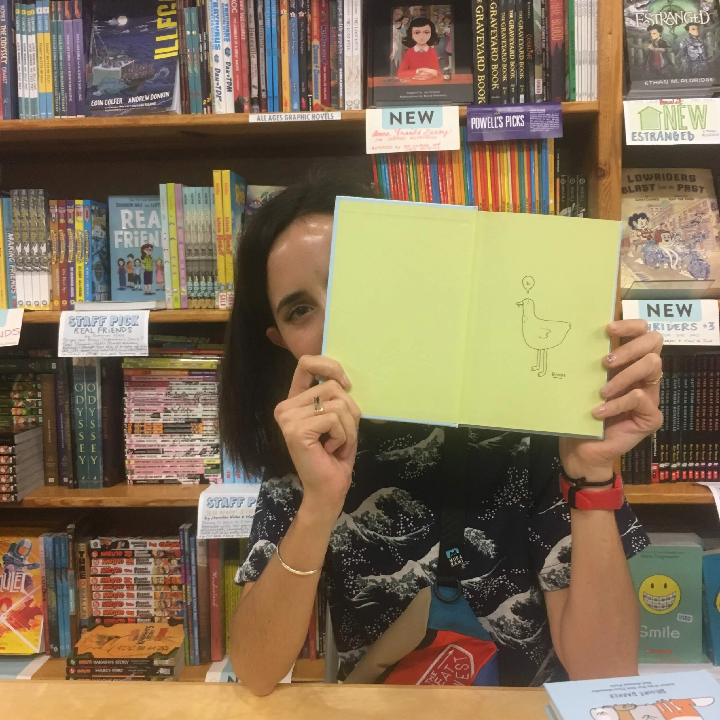 Person holding open a book with a duck illustration, standing in front of a bookshelf filled with graphic novels and books.
