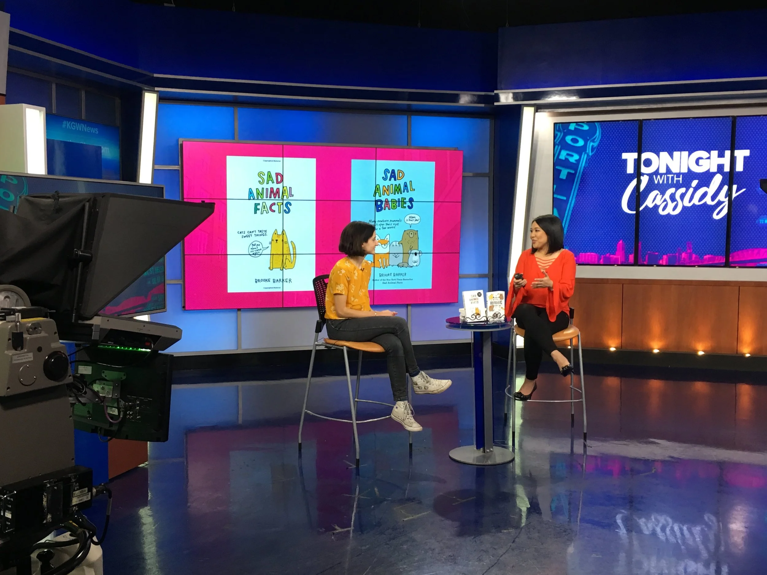 Two women sitting on chairs in a TV studio, with a display showing book covers 'Sad Animal Facts' and 'Sad Animal Babies'. Studio lighting and cameras visible.