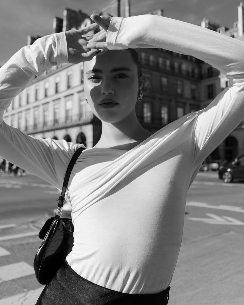 Black and white photo of a woman with arms raised above her head, standing on a city street with buildings in the background.