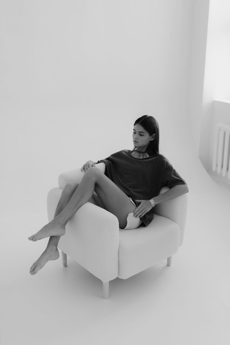 Black and white photo of a woman sitting in a white armchair in a minimalist room, with her legs crossed and her gaze downward.