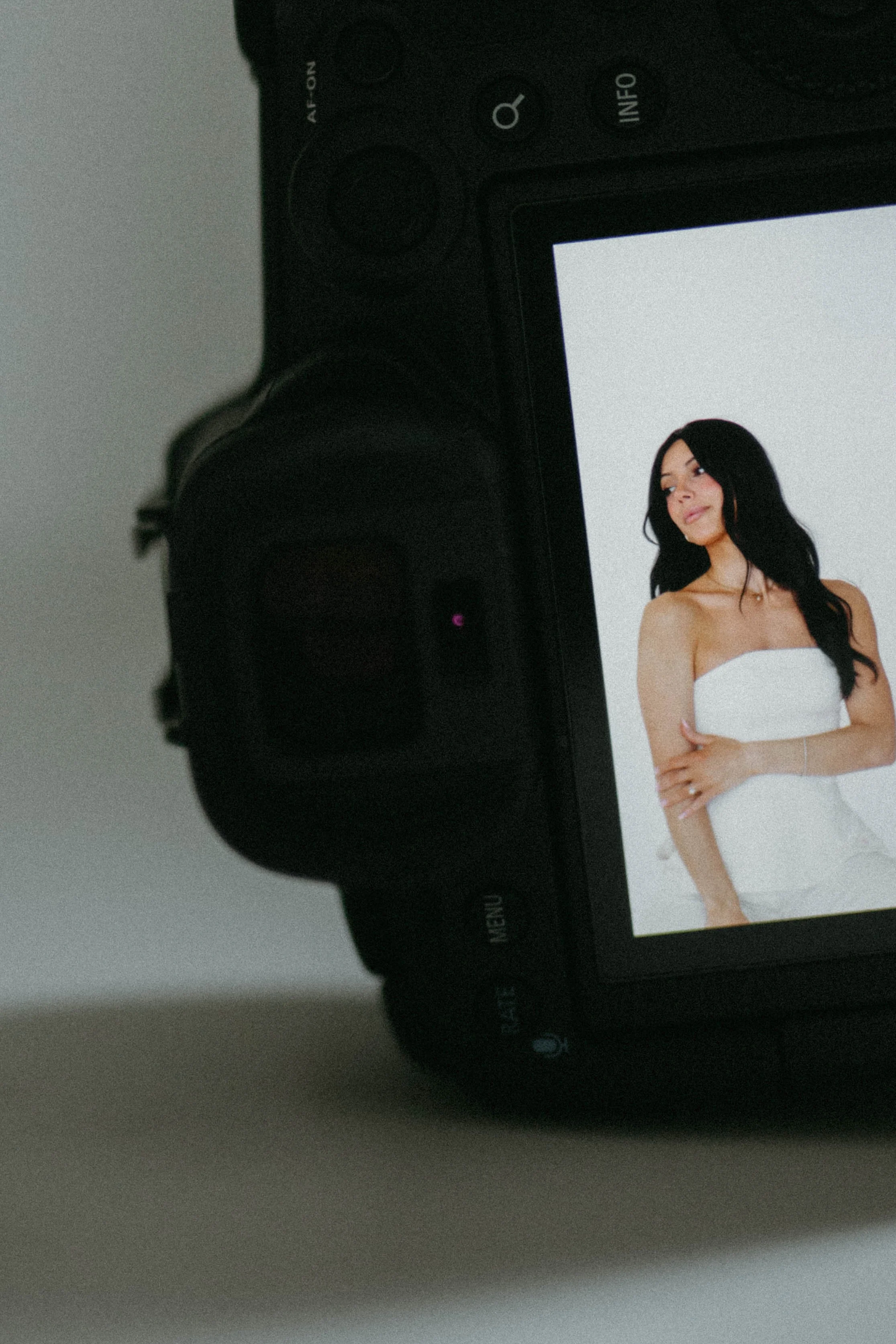 Digital camera screen showing a woman with black hair in a white strapless dress, posing against a white background.