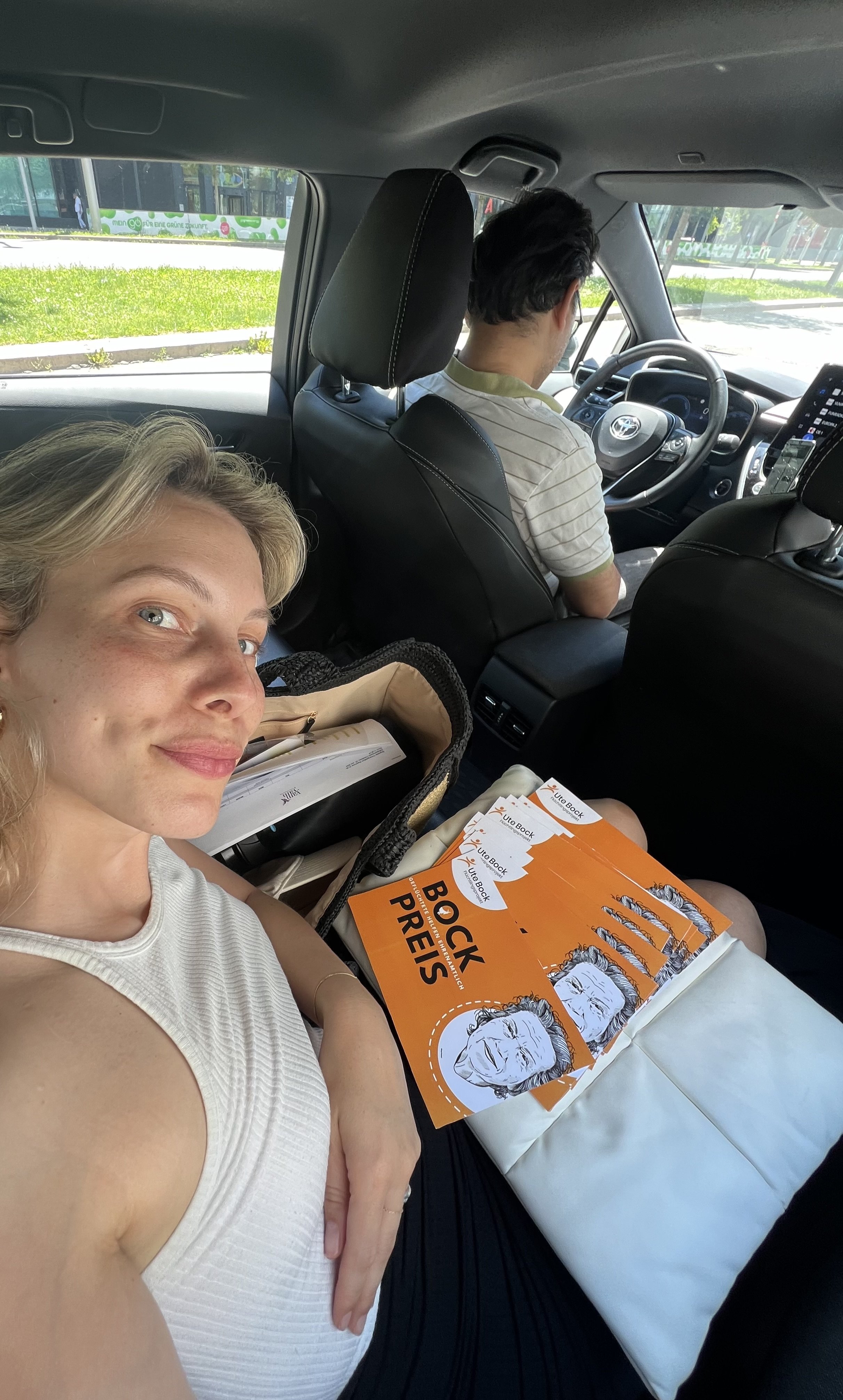 Eine Frau liegt auf einem Sitz im Auto, hält ein Buch mit der Aufschrift 'Bock Preis' und sitzt neben einem Mann, der das Auto fährt.