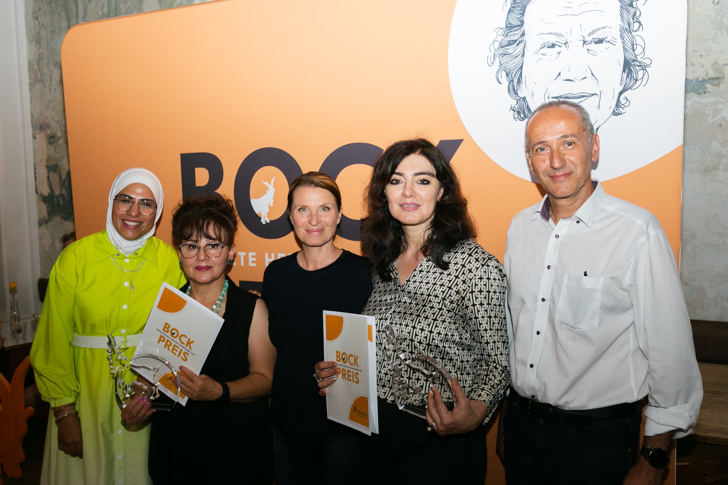 Fünf Menschen bei einer Preisverleihung vor einer orangefarbenen Hintergrund mit einem weißen Gesicht und Text "BOCK".