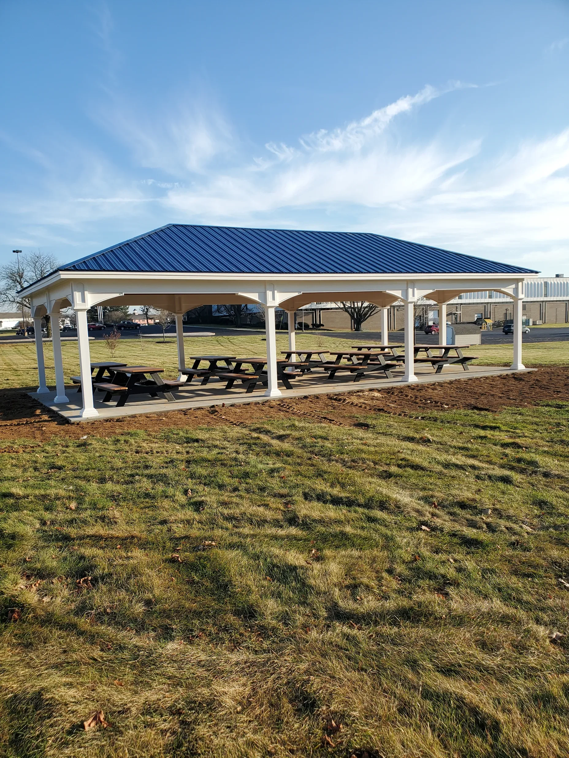 A park shelter with a blue metal roof and white support columns, surrounded by a grassy area and outdoor picnic tables.