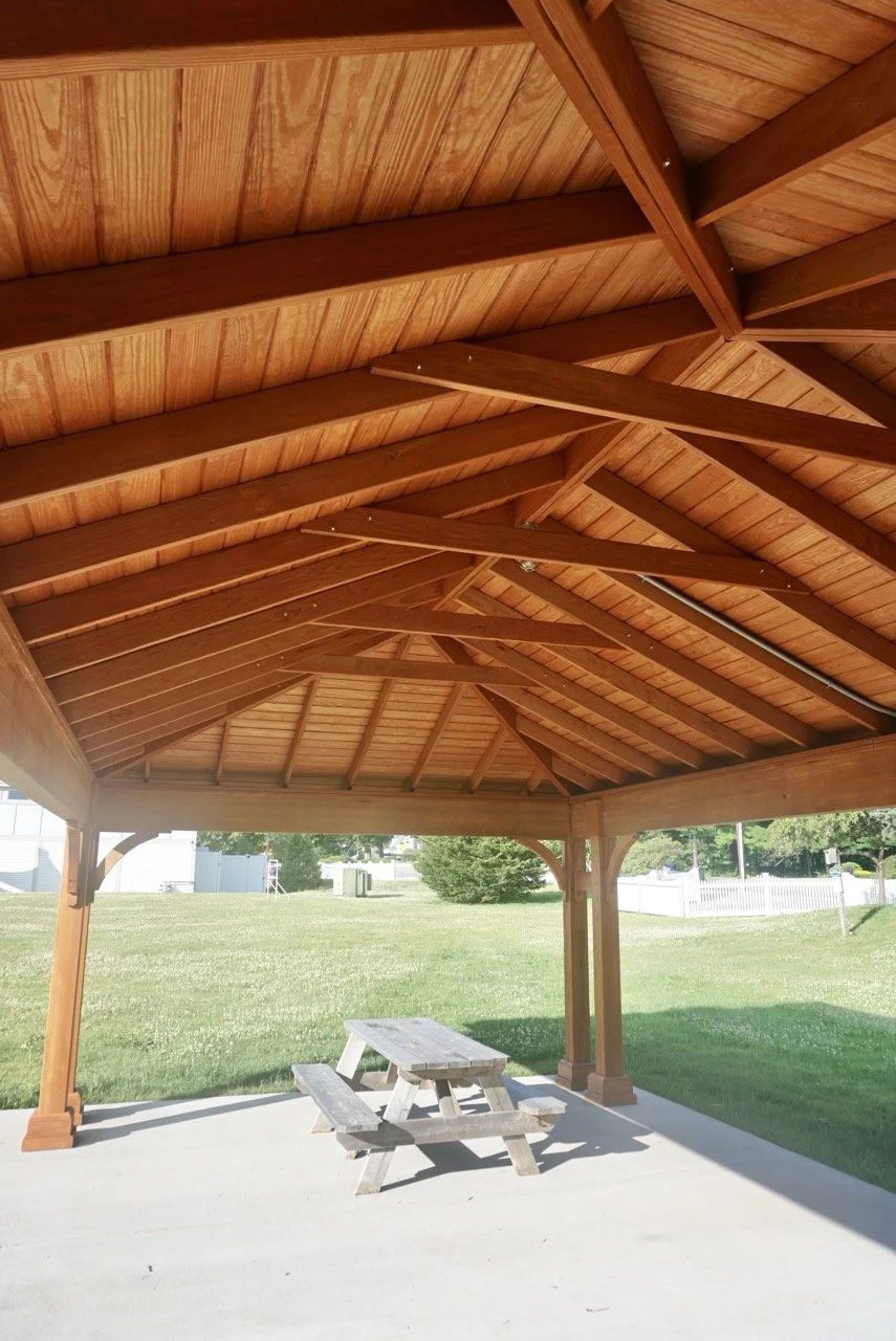 Wooden pavilion with a picnic table underneath, situated on a concrete slab in a grassy outdoor area.