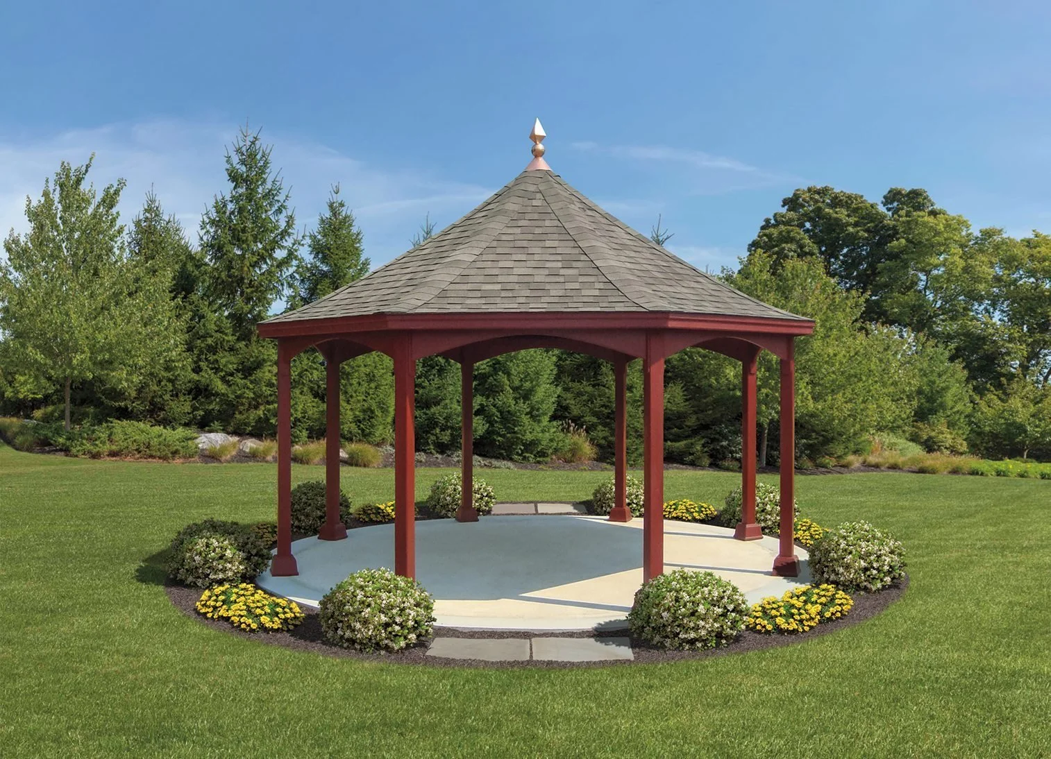 A red wooden gazebo with a shingled roof, situated on a circular concrete patio, surrounded by landscaped flower beds with yellow and white flowers, in a grassy park with trees and blue sky in the background.