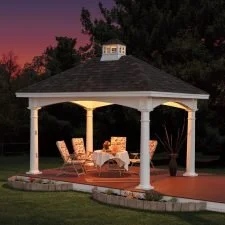 A white gazebo with a dark shingled roof, situated on a paved patio with outdoor furniture, surrounded by greenery and trees at sunset.