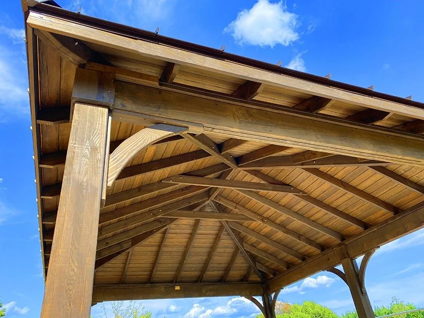 Close-up of a wooden pavilion roof structure with blue sky and clouds in the background.