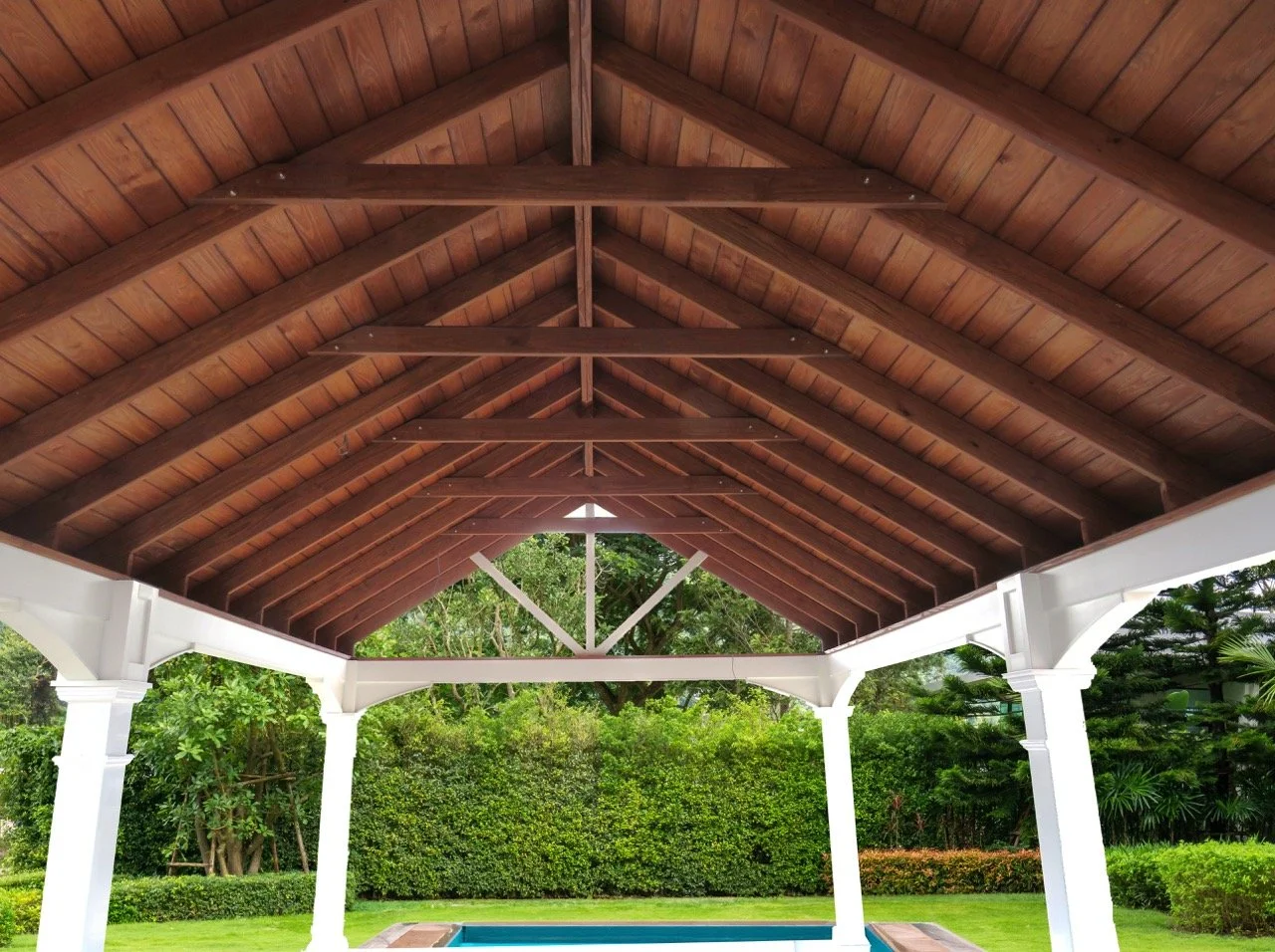 View of a covered outdoor patio with a wooden roof and white columns, overlooking a backyard with green grass, bushes, and trees.