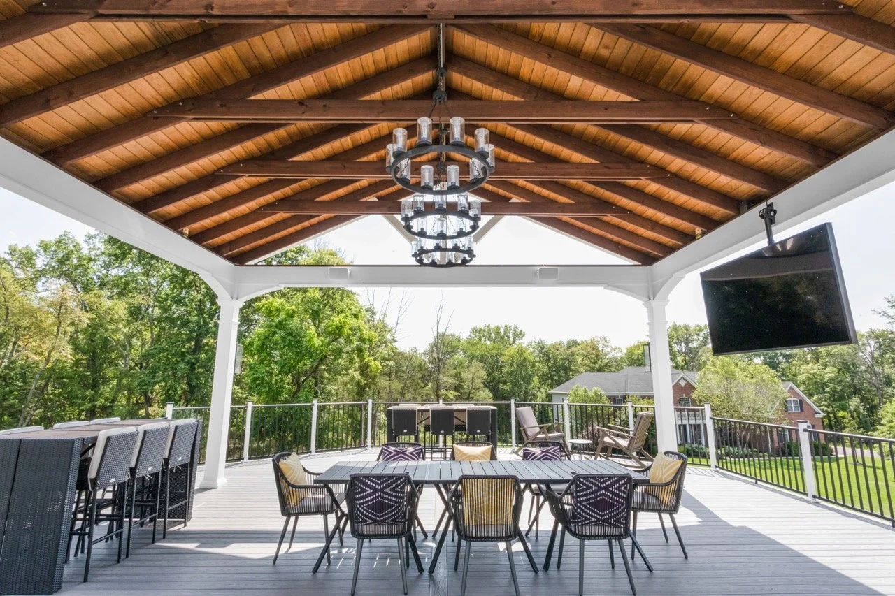 An outdoor covered patio with a wooden ceiling, a chandelier, a dining table with chairs, and a large flat-screen TV mounted on the ceiling
