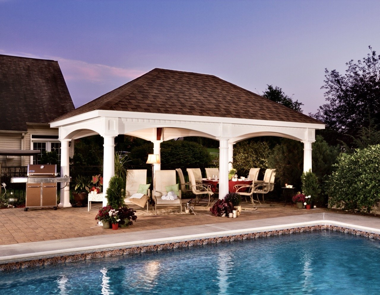 A backyard patio with a pool, a white pavilion with columns, outdoor seating, a grill, potted plants, and decorative flowers, during twilight.