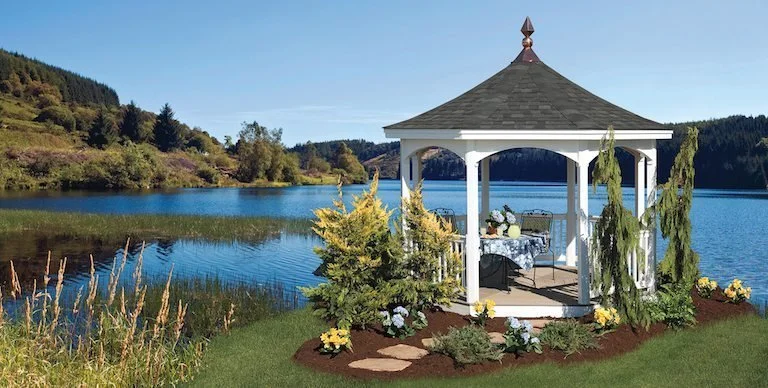 A white gazebo with a dark gray roof on a grassy lawn near a lake, surrounded by green plants and yellow flowers, with trees and hills in the background.