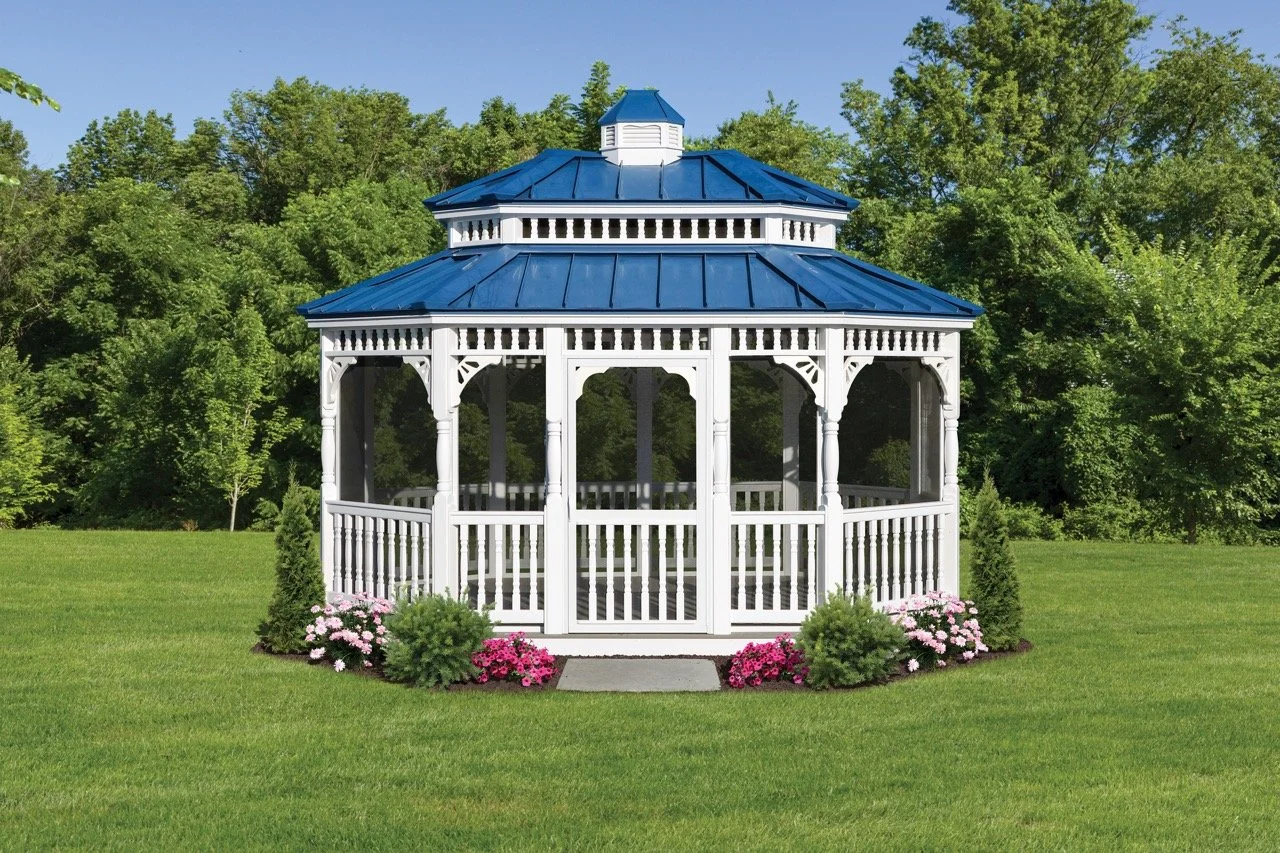White pavilion with blue roof in a grassy yard, surrounded by pink and green bushes and trees.