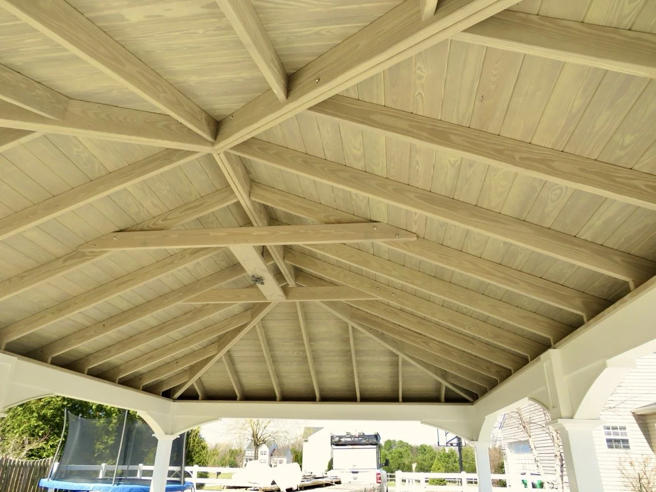 View of a wooden roof structure with exposed beams and a ceiling, open on the sides, with outdoor scenery including a trampoline and a house in the background.