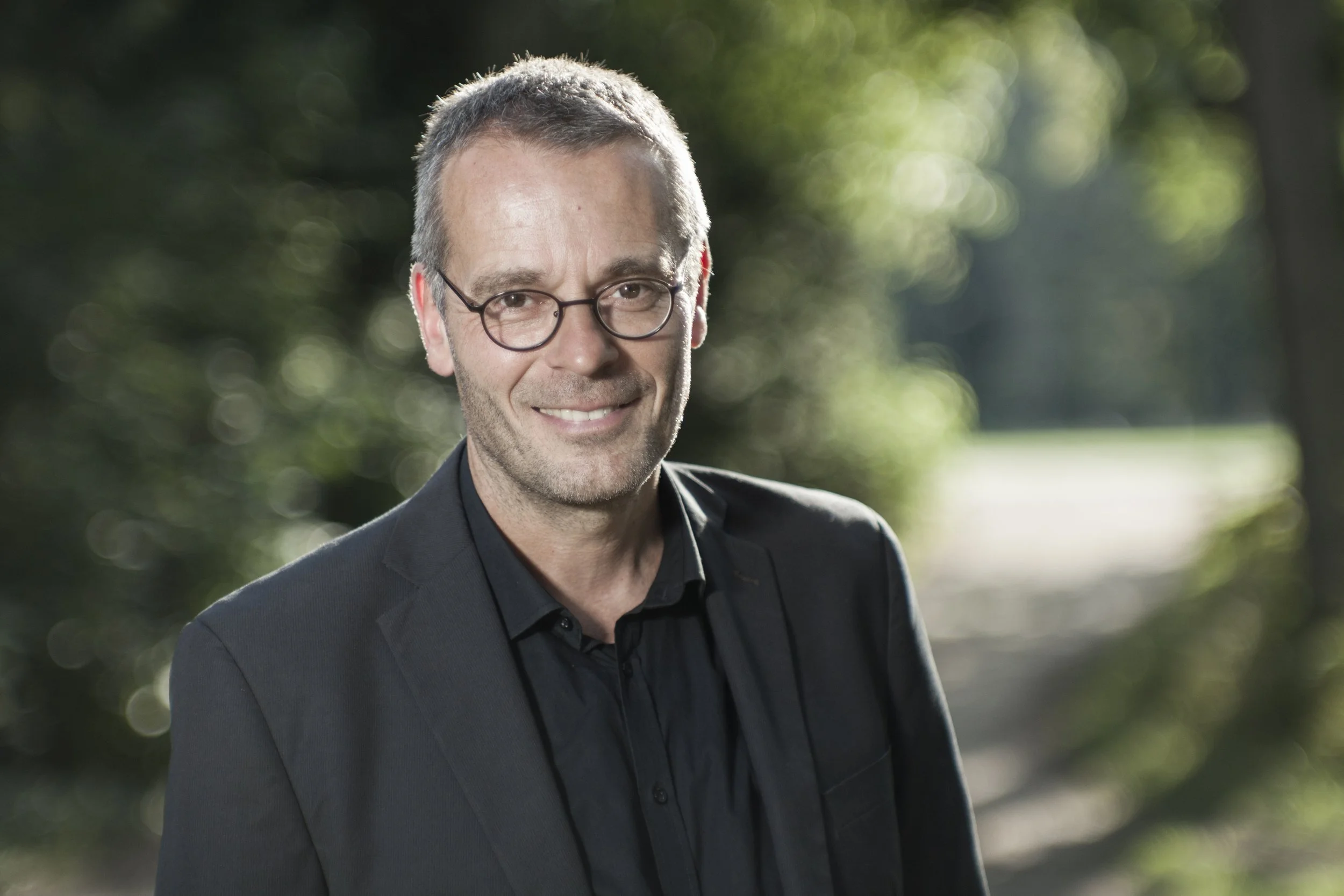 Andreas Ulich mit Brille trägt einen schwarzen Anzug und steht im Freien vor einem grünen, bewaldeten Hintergrund.